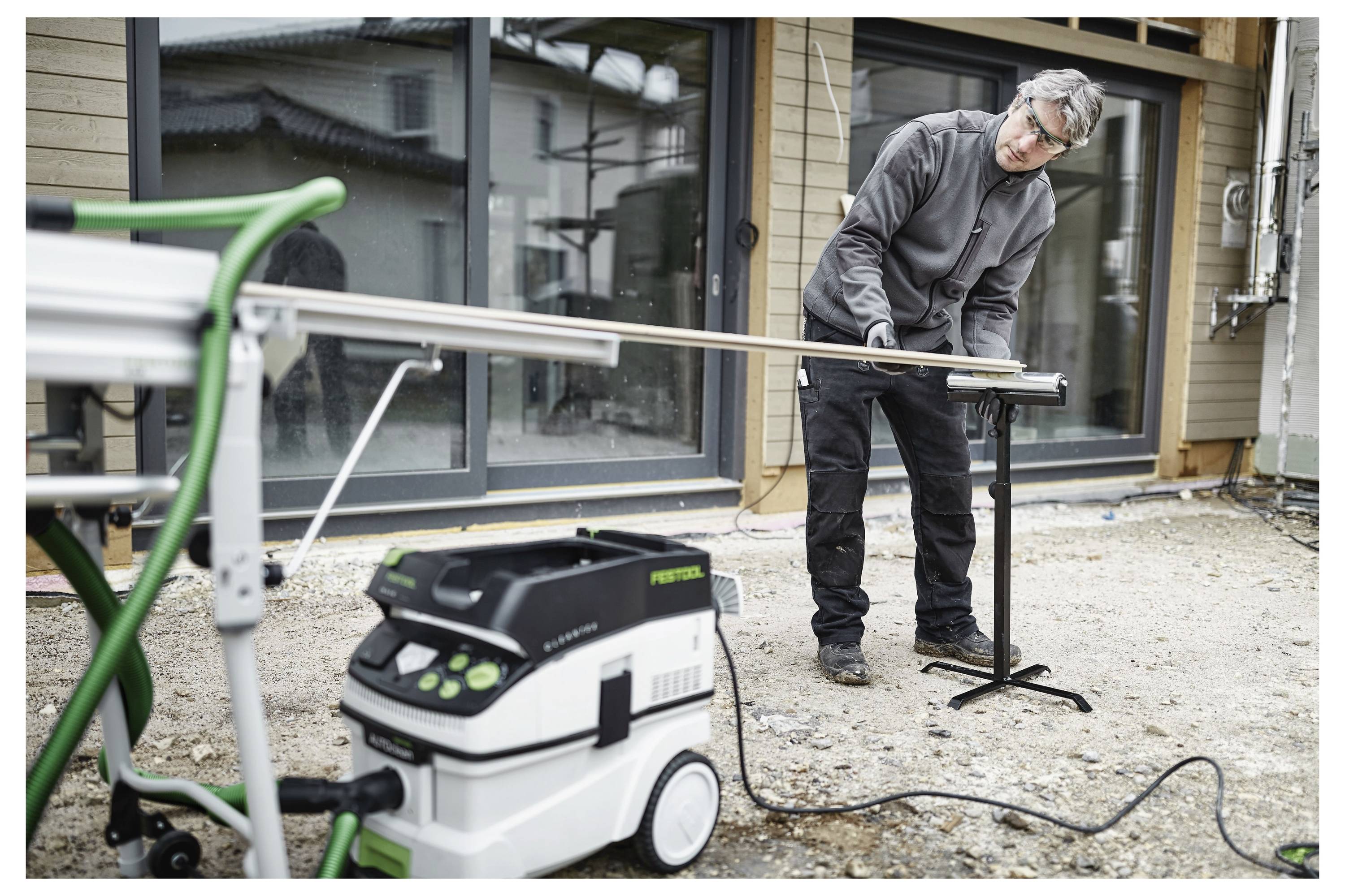 Eine Person schneidet auf einer Baustelle ein langes Stück Holz mit einer Säge und einem Staubsauger zu, während sie vor einem renovierungsbedürftigen Gebäude steht.