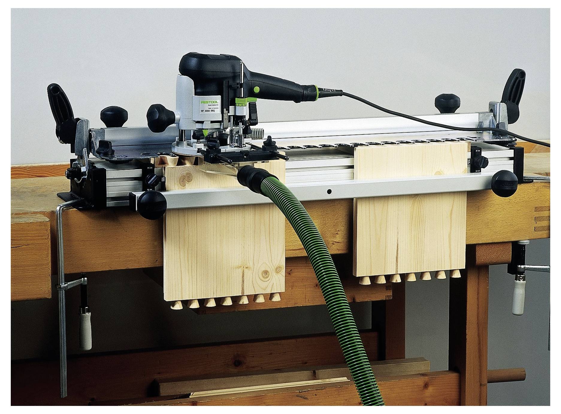 Ein Holzbearbeitungs-Schwalbenschwanzlehre auf einer Werkbank, die zum präzisen Schneiden von Verbindungen verwendet wird. Sie ist mit einem Elektrowerkzeug und einem grünen Absaugschlauch ausgestattet.