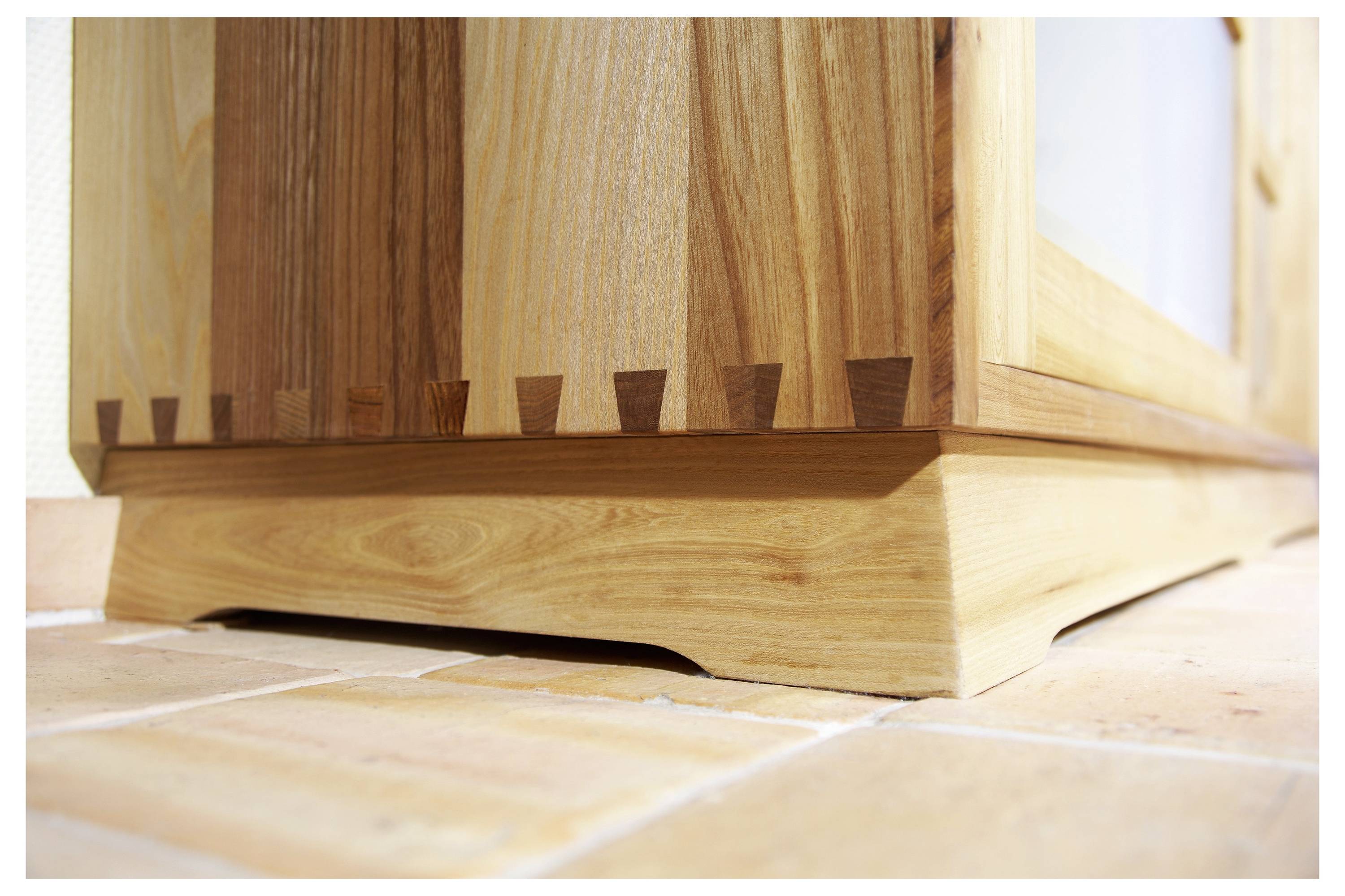 Ein hölzerner Schrank steht auf einem gefliesten Boden und zeichnet sich durch Schwalbenschwanzzapfen aus, die die Seitenpaneele mit der Basis verbinden und die handwerkliche Kunst hervorheben.