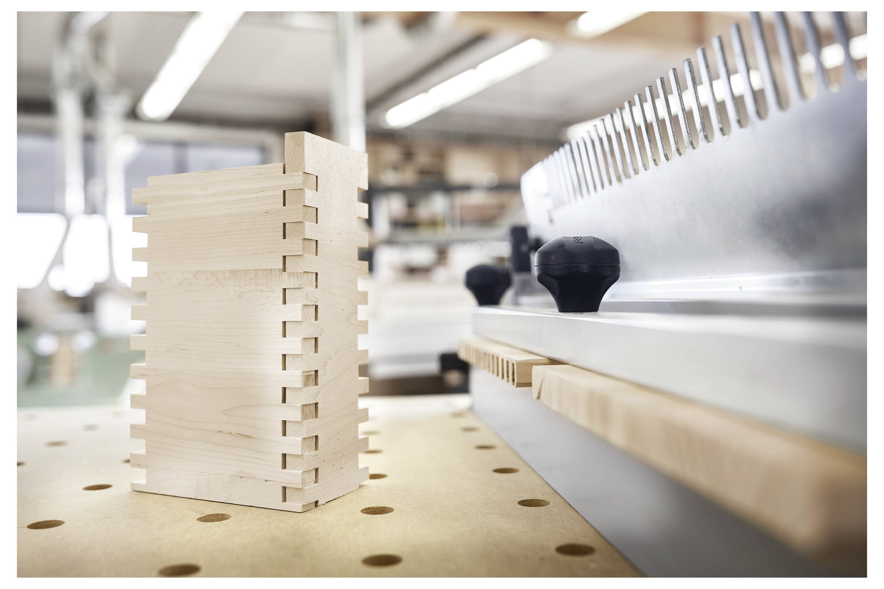 Eine Holzkiste mit Schwalbenschwanzverzapfungen sitzt auf einer perforierten Werkbank in einer Schreinerwerkstatt, neben einem Metallwerkzeug mit Drehreglern.