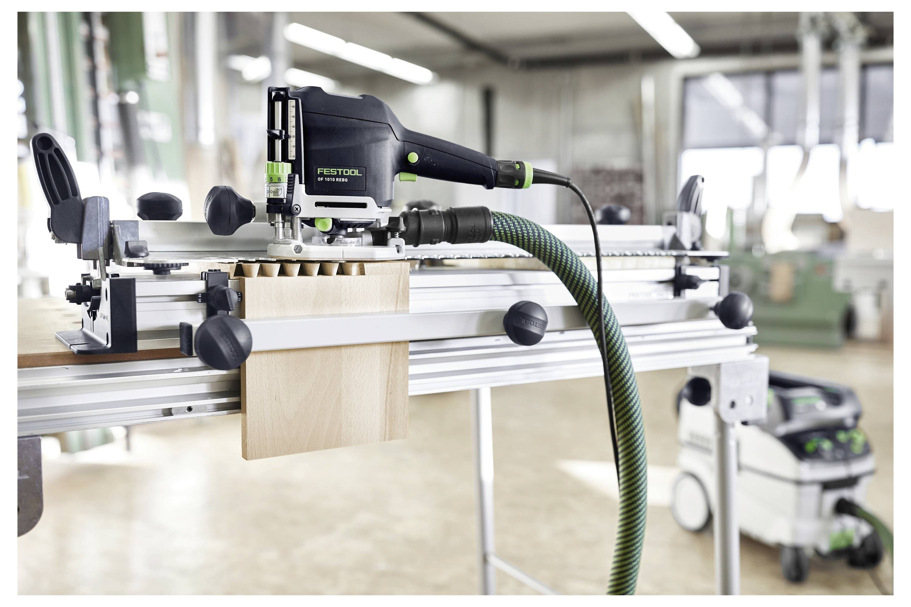 Werkstattszene mit einem auf einer Werkbank montierten Festool-Werkzeug, das offenbar für Holzbearbeitung verwendet wird, mit angeschlossenen Zubehörteilen und einem Industriestaubsauger im Hintergrund.