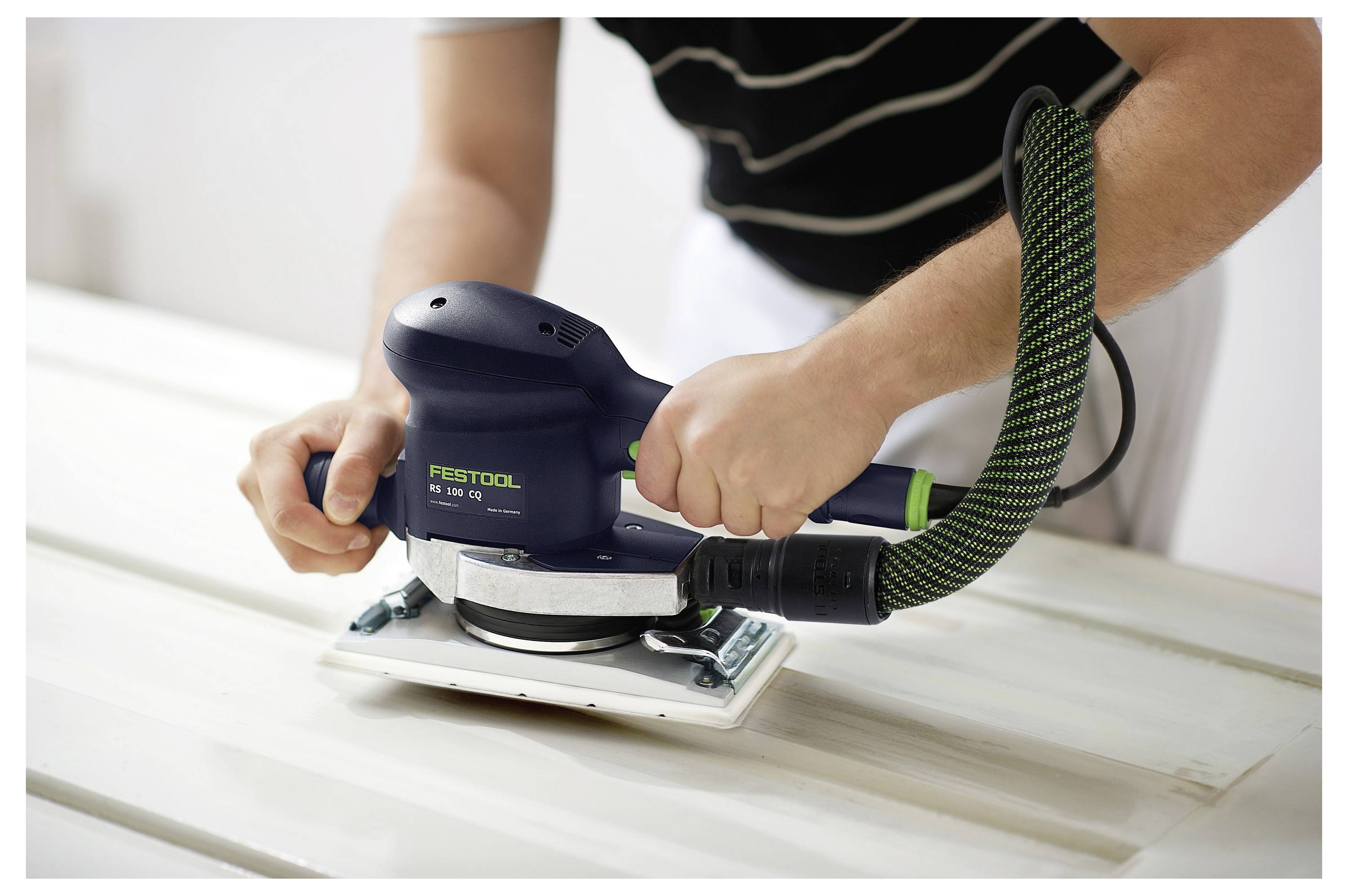 Eine Person verwendet einen Elektroschleiffer, um eine Holzoberfläche zu glätten, und hält das Gerät mit beiden Händen. Der Schleiffer ist mit einem Schlauch verbunden.