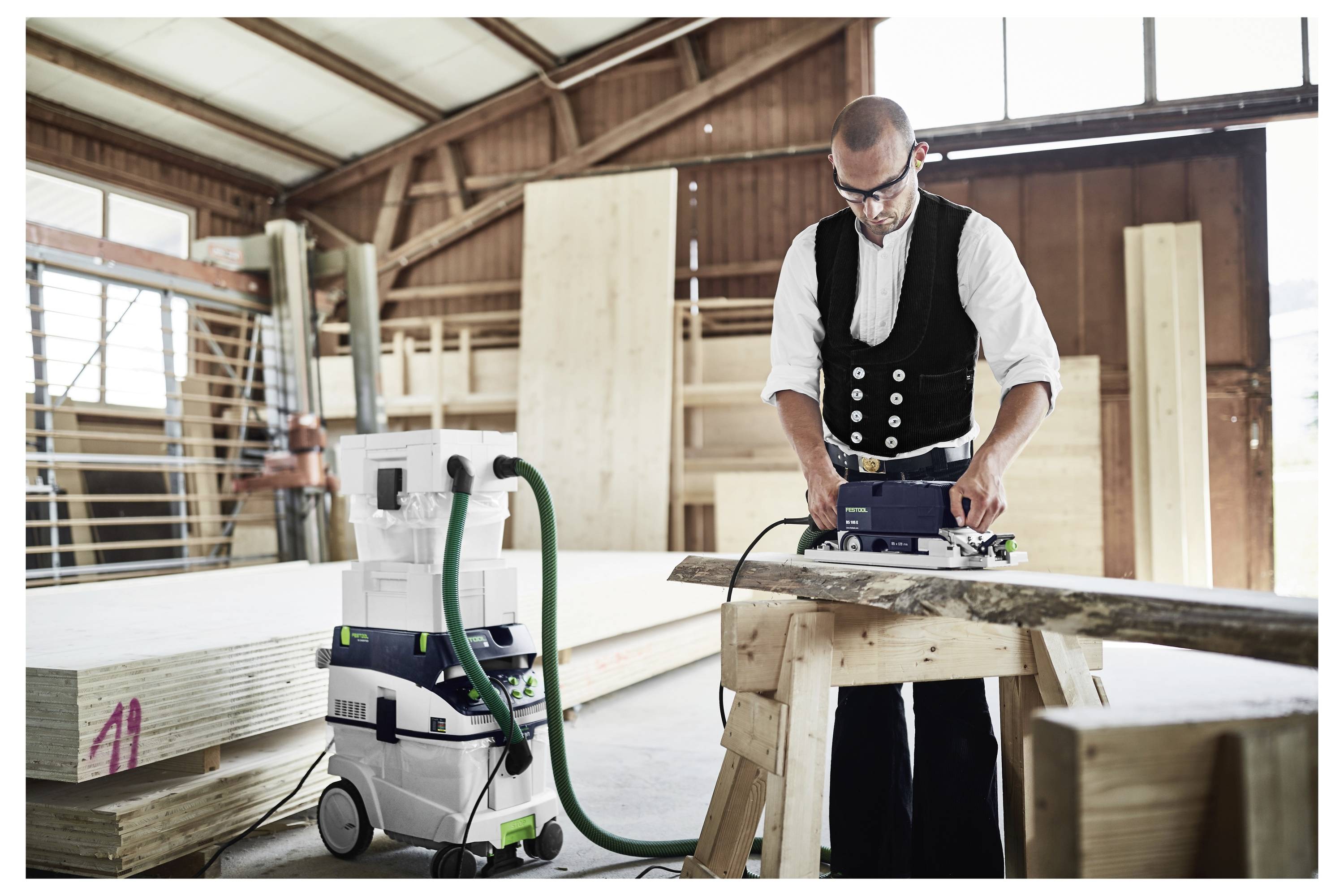 Eine Person nutzt ein Elektrowerkzeug, um Holz in einer Werkstatt zu schneiden, trägt Schutzbrille und traditionelle Kleidung. Ein Staubsauger ist an das Werkzeug angeschlossen.