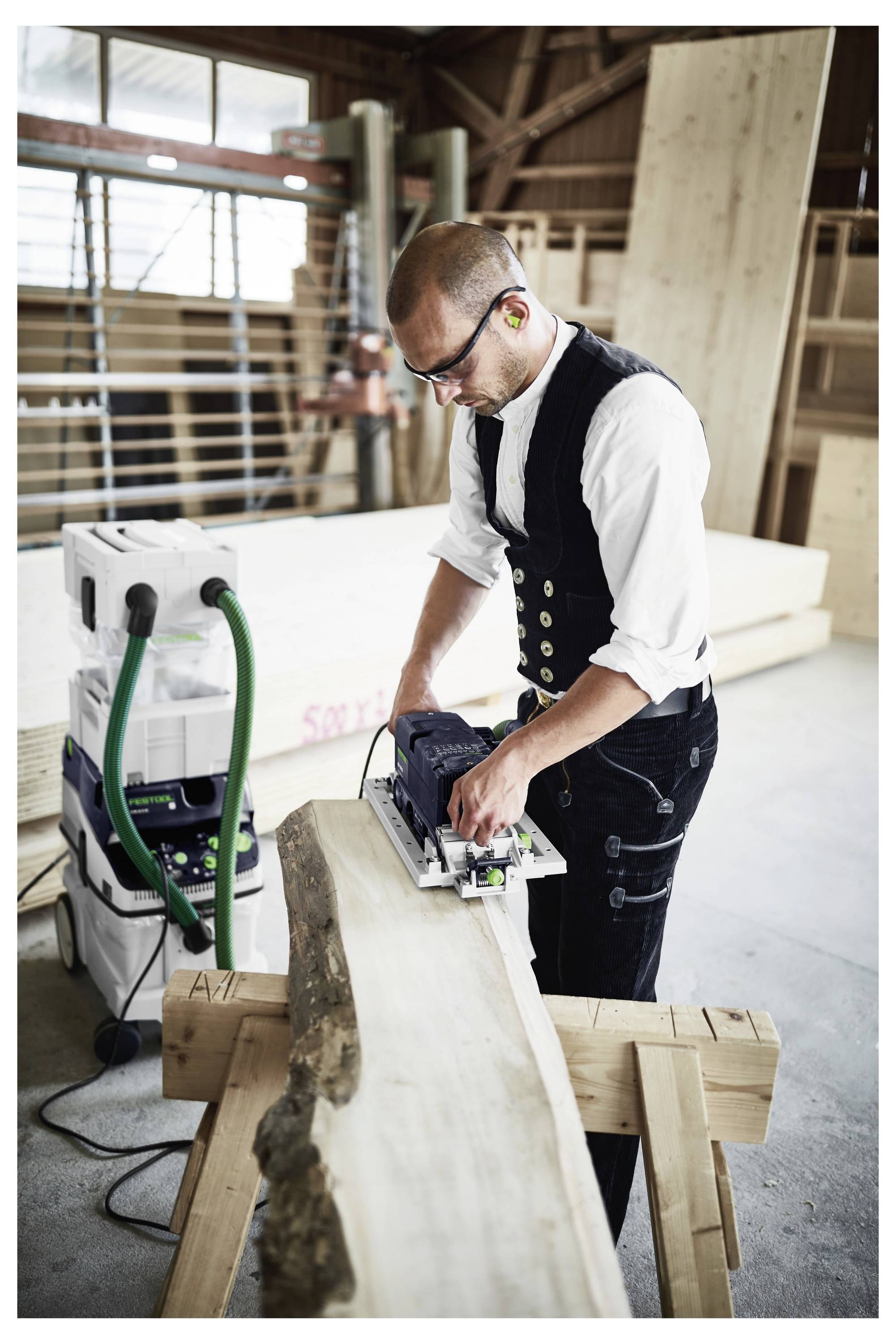 Ein Handwerker in einer Werkstatt nutzt ein Elektrowerkzeug, um ein großes Stück Holz auf einem Sägebock zu zuschneiden, trägt Schutzausrüstung und konzentriert sich auf seine Aufgabe.