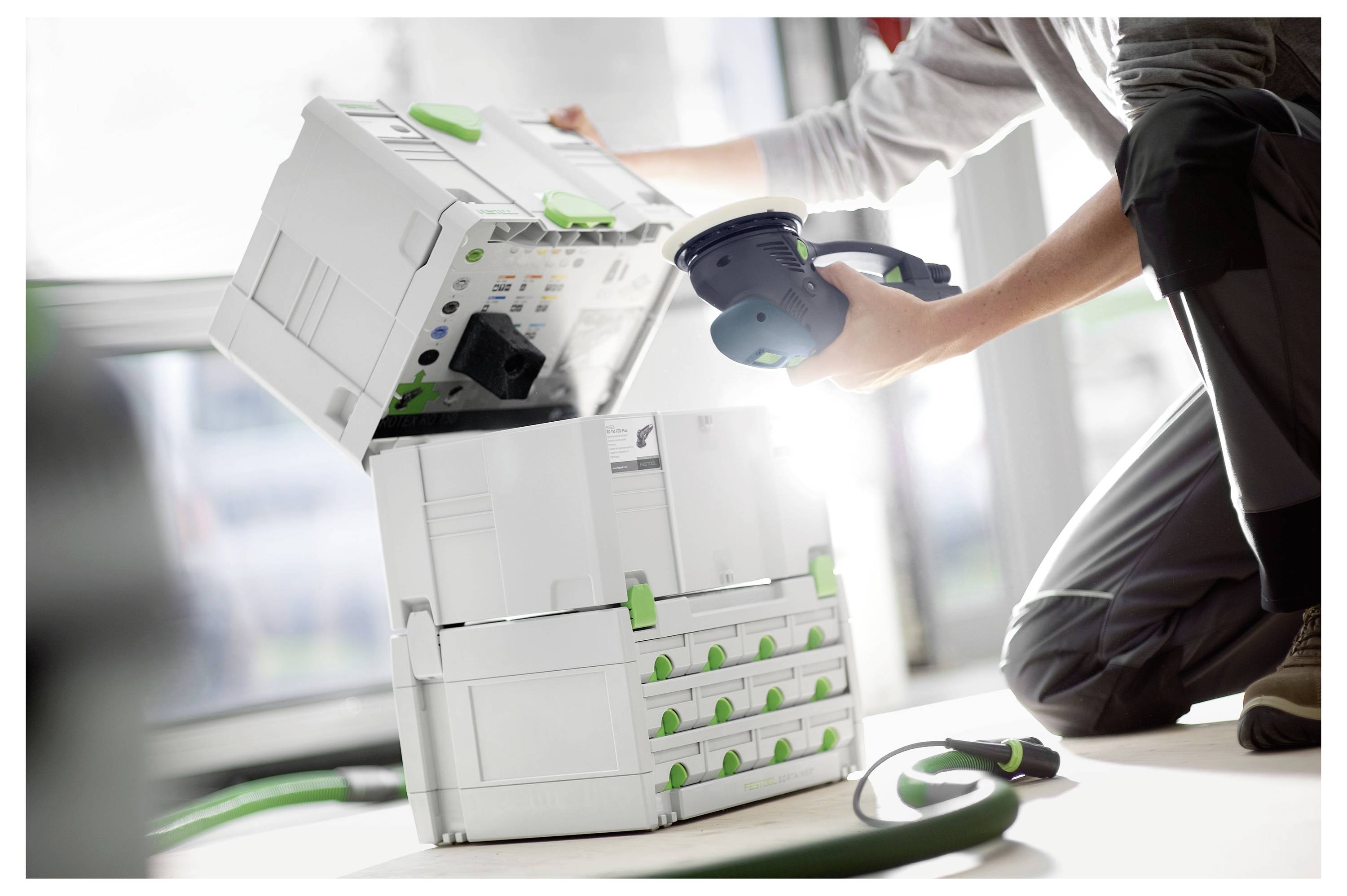 Eine Person, die ein Werkzeug handhabt, mit einem weißen und grünen modularen Lagersystem in der Nähe, wobei Ordnung und Effizienz in einer Werkstattumgebung hervorgehoben werden.