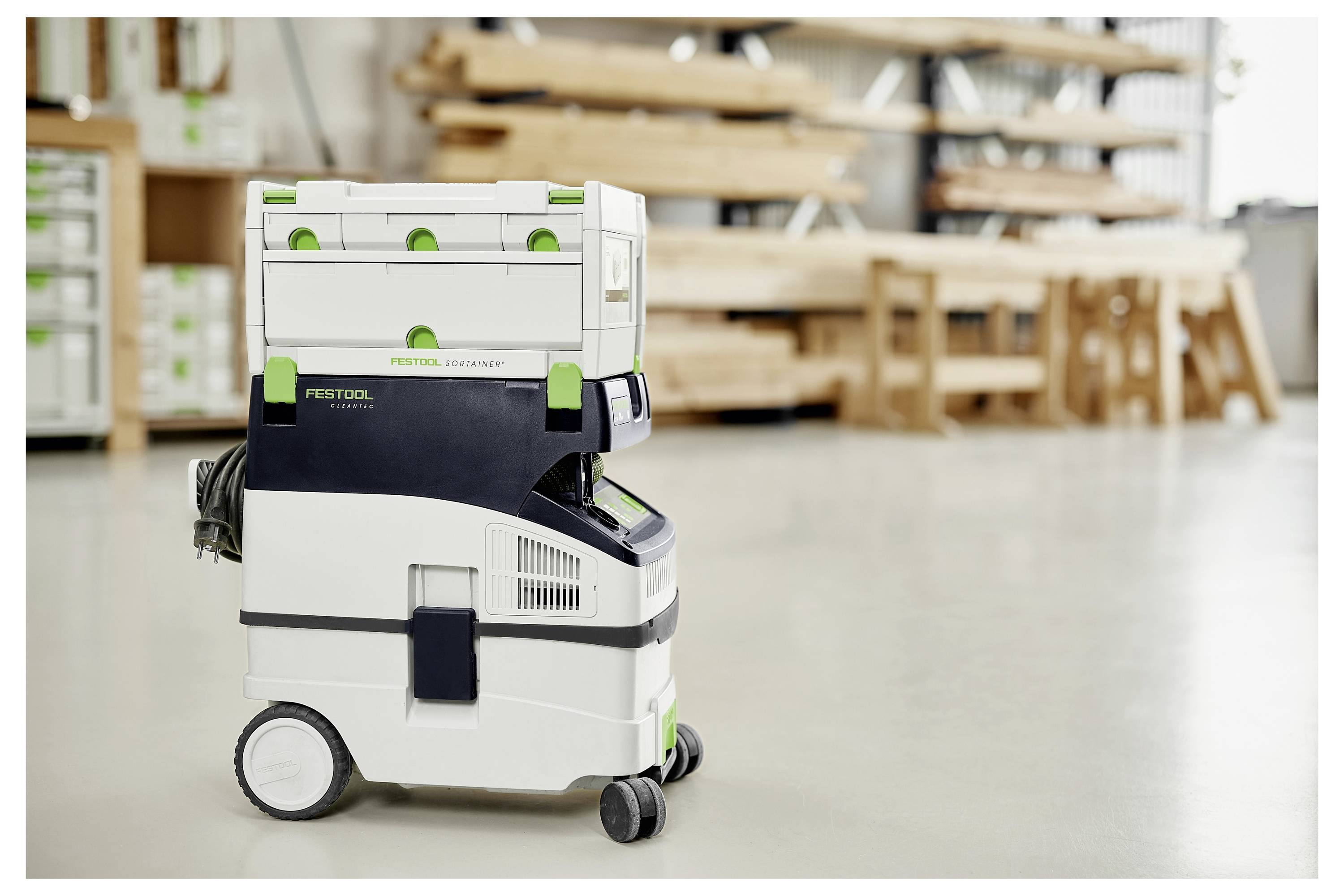 Ein Festool-Staubabsauger steht auf dem Boden einer Holzwerkstatt, mit unscharfen Werkbänken aus Holz und Regalen im Hintergrund.