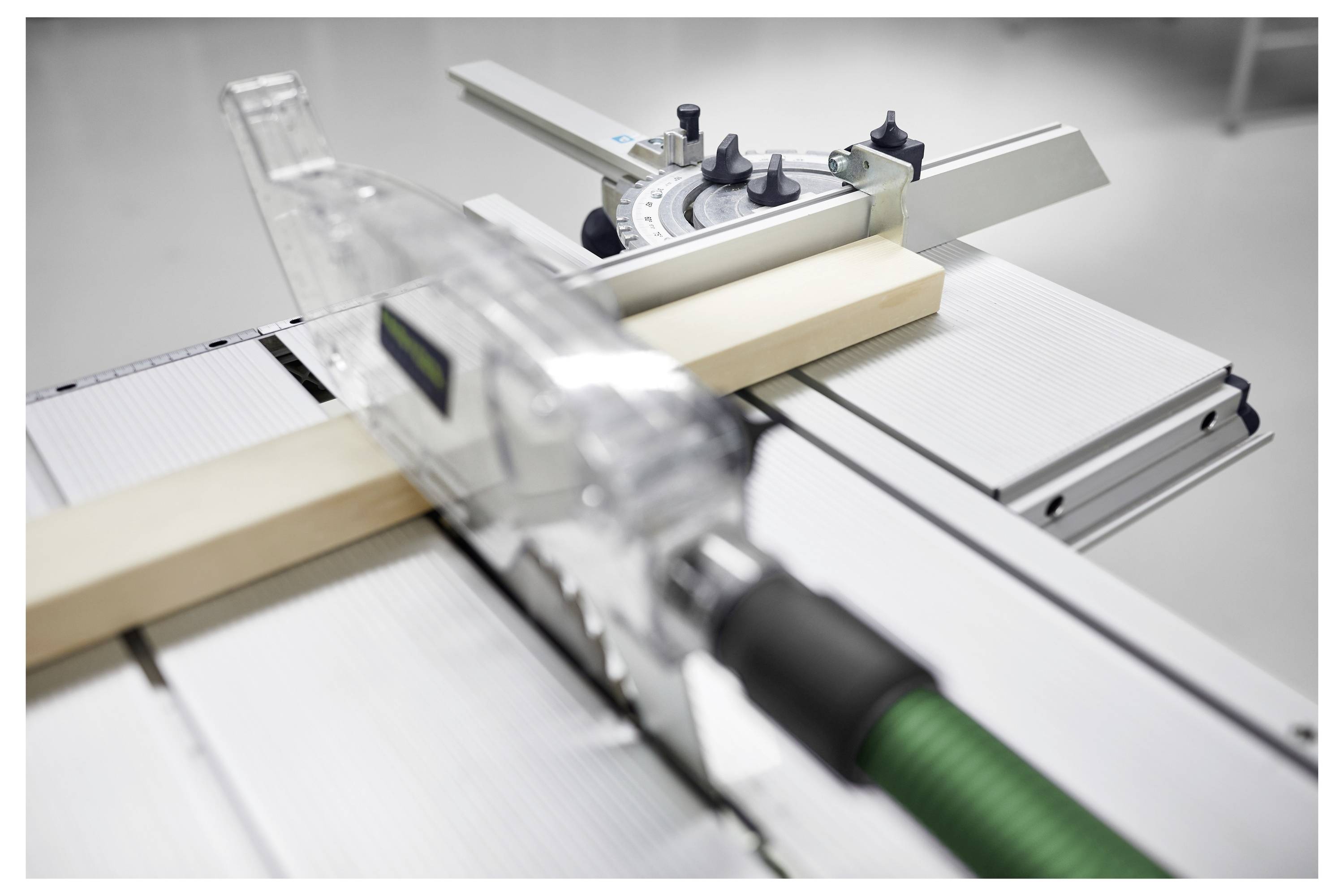 Eine Tischkreissäge mit einem Holzstück auf ihrer Oberfläche. Das Sägeblatt ist teilweise sichtbar, und ein grüner Schlauch ist an der Seite angeschlossen.