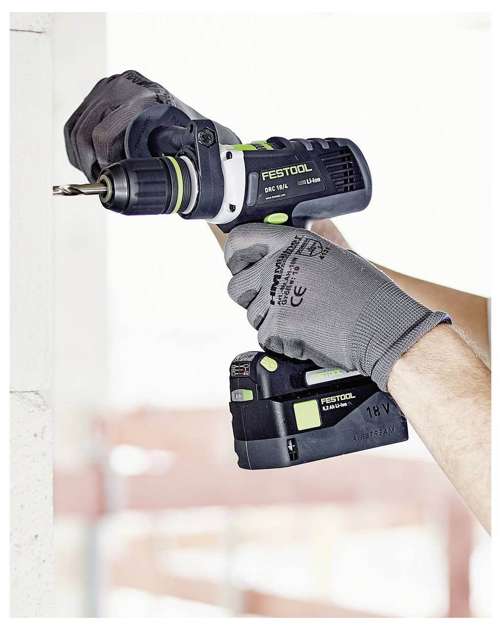 Eine Person, die Handschuhe trägt, verwendet einen Festool-Bohrer, um ein Loch in eine weiße Wand zu bohren, was auf Bau- oder Reparaturarbeiten hindeutet.