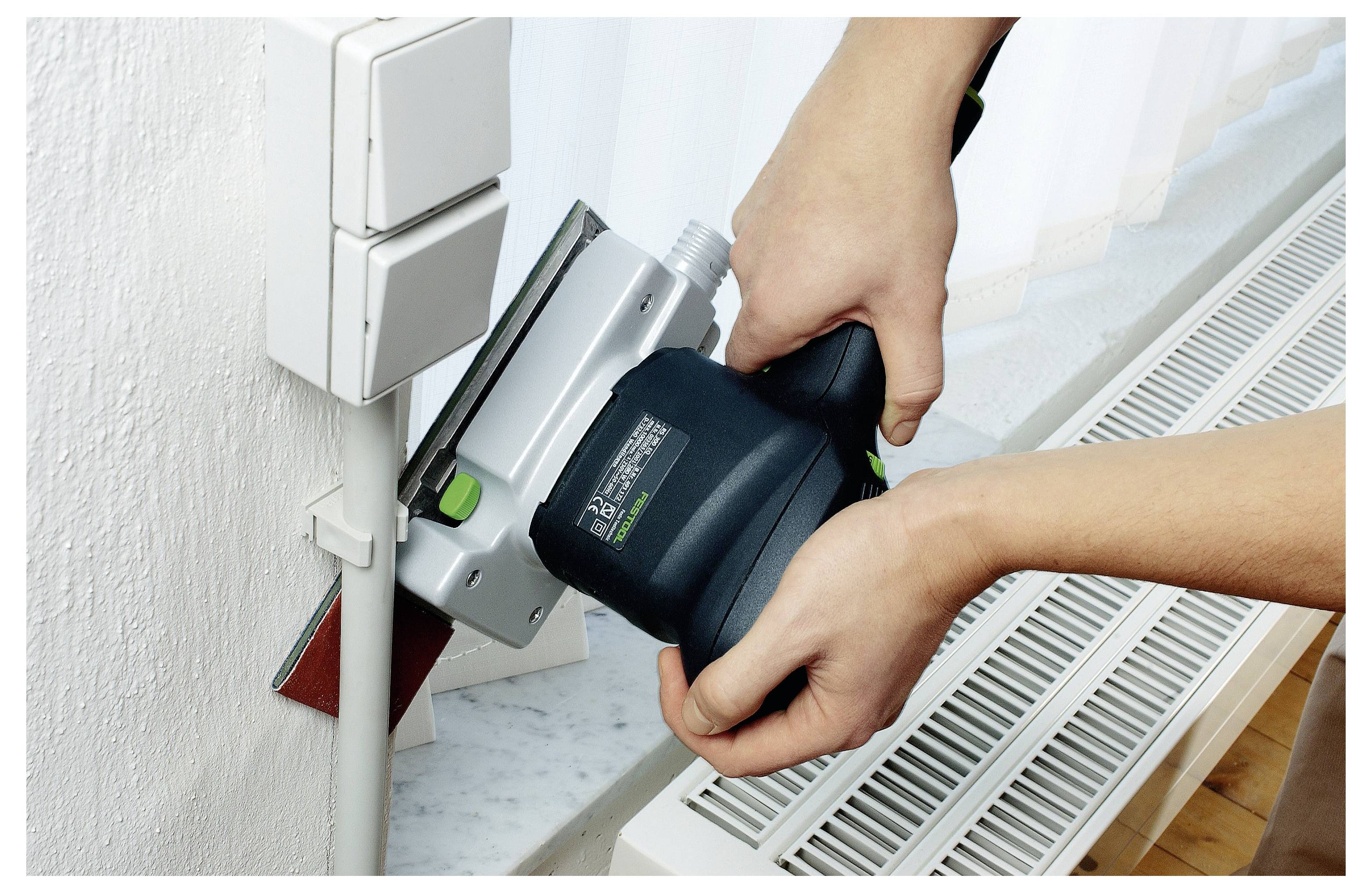 Zwei Hände benutzen einen Elektroschleiffer auf einer Fensterbank neben einem Heizkörper und glätten die Oberfläche für gleichmäßigkeit.