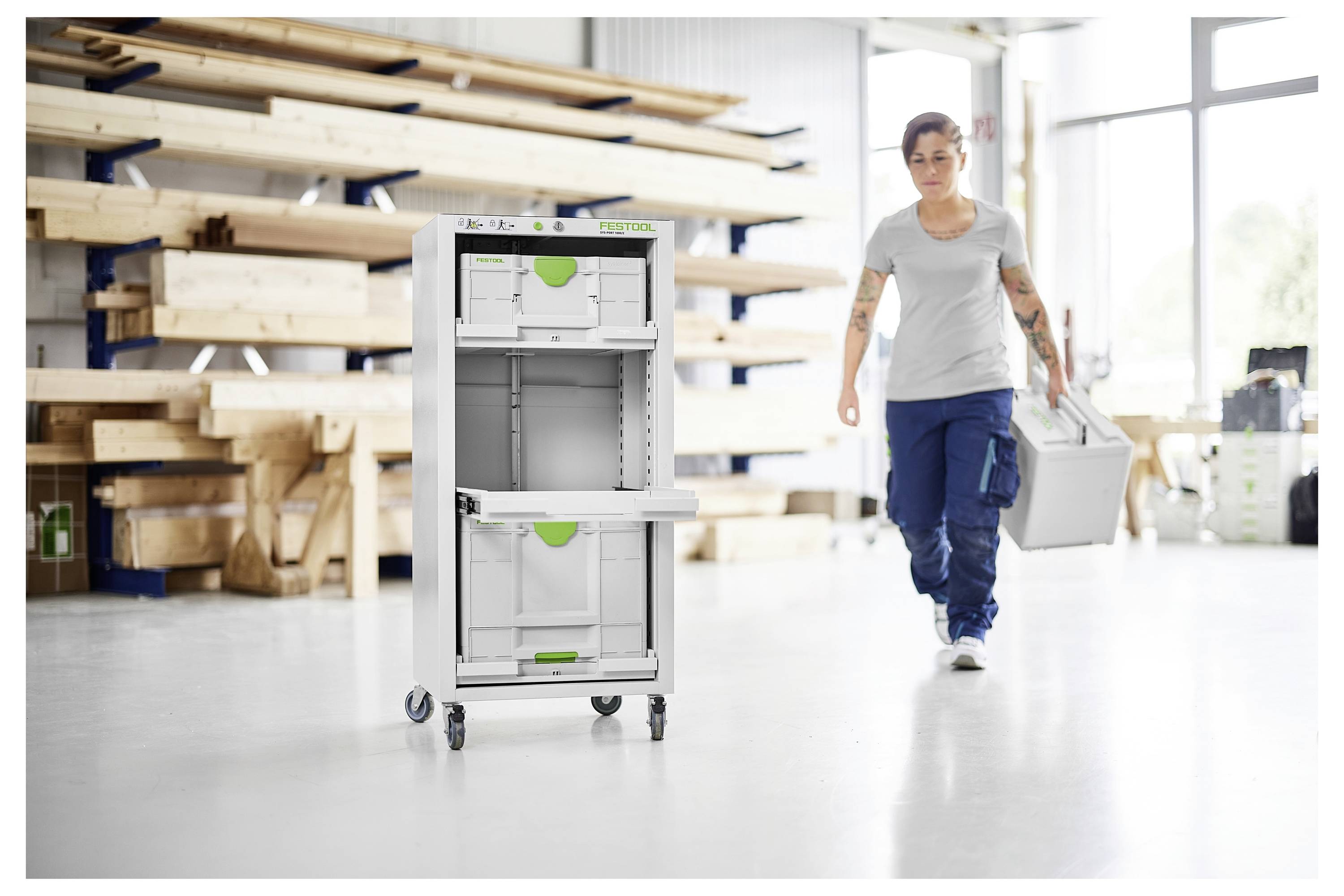 Eine Person in einer Werkstatt trägt eine Lagerkiste zu einer mobilen Lagereinheit mit offenen Schubladen. Holzbalken sind im Hintergrund gestapelt.