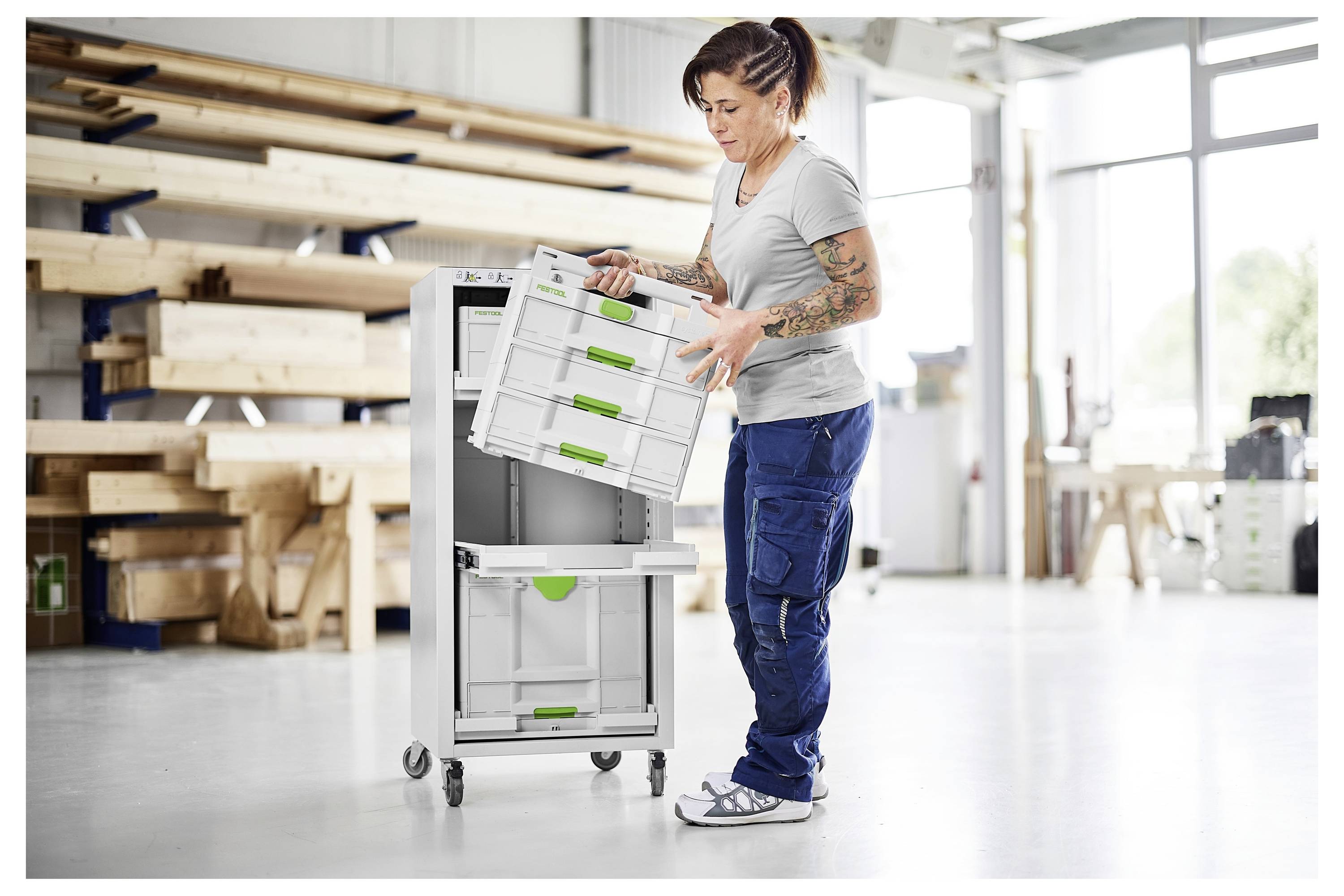 Eine Person in einer Werkstatt stellt eine Schublade in eine hohe, fahrbaren Lagerbehälter. Im Hintergrund sind Regale und große Fenster sichtbar.