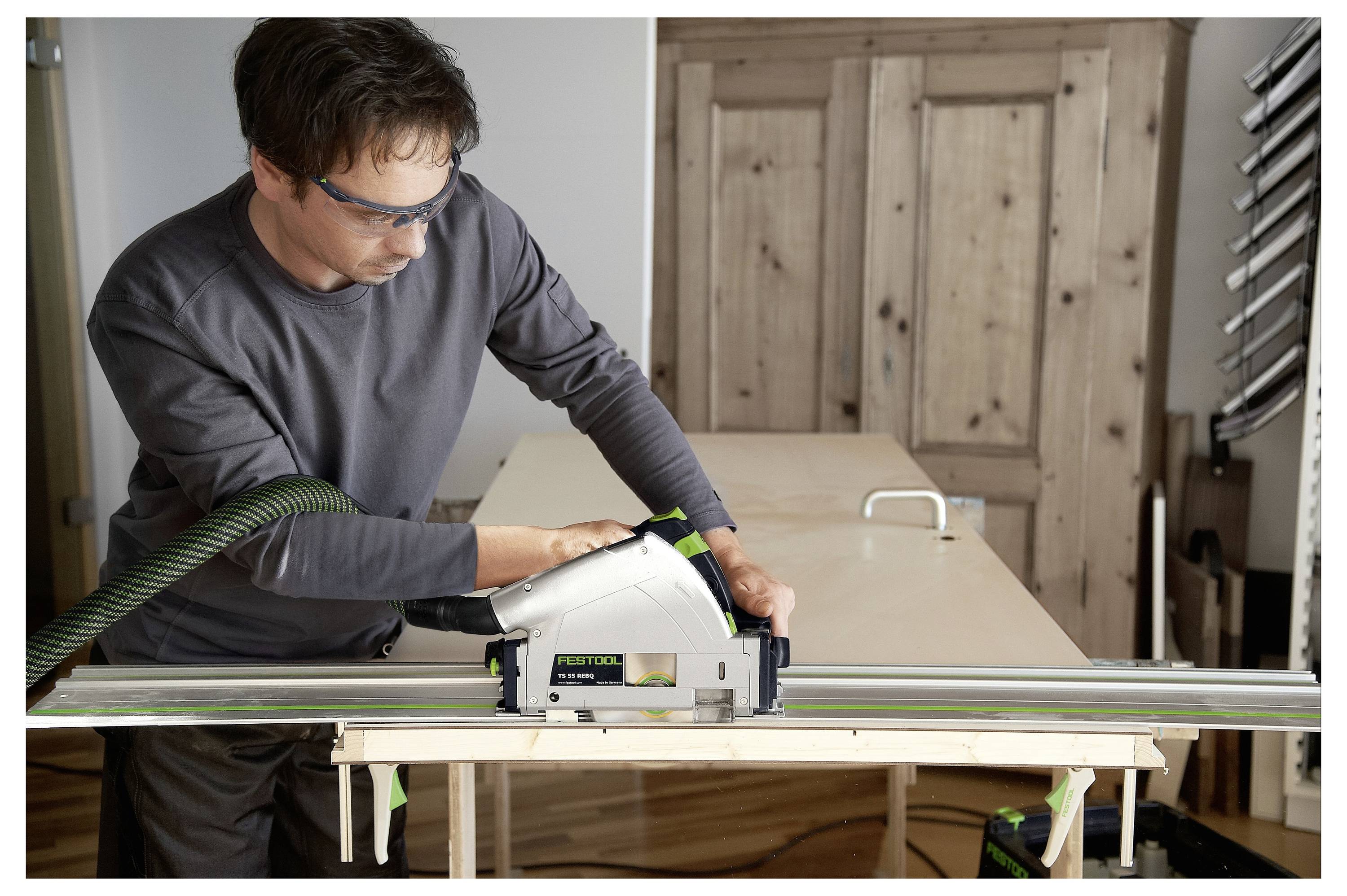 Eine Person, die eine Schutzbrille trägt, verwendet eine Tauchkreissäge, um ein großes Holzblatt auf einem Tisch in einer Werkstattumgebung zu schneiden.