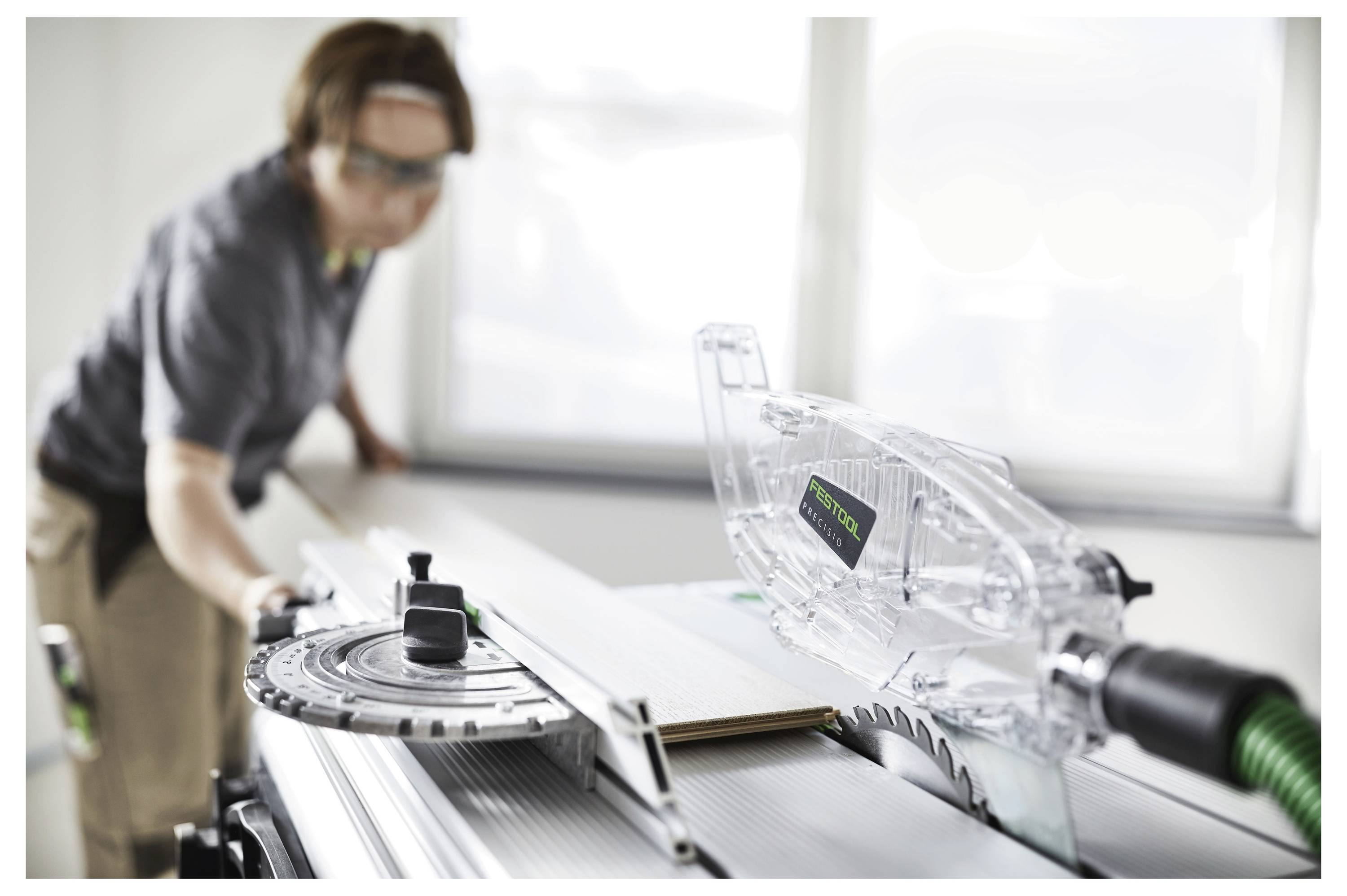 Eine Person mit Schutzbrille bedient eine Tischkreissäge, um ein Holzstück zu schneiden, wobei der Fokus auf der klaren Schutzvorrichtung der Säge liegt.