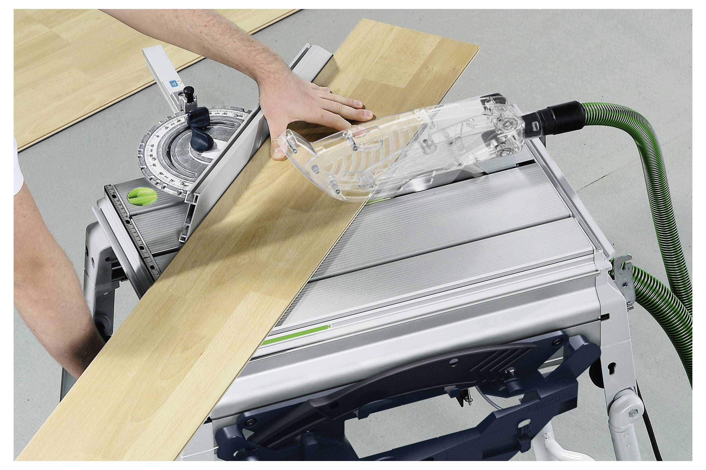 Eine Person führt ein Holzbrett durch eine Tischkreissäge mit einem durchsichtigen Schutzvorrichtung, um einen präzisen Schnitt zu gewährleisten. Sicherheitsausrüstung ist sichtbar.