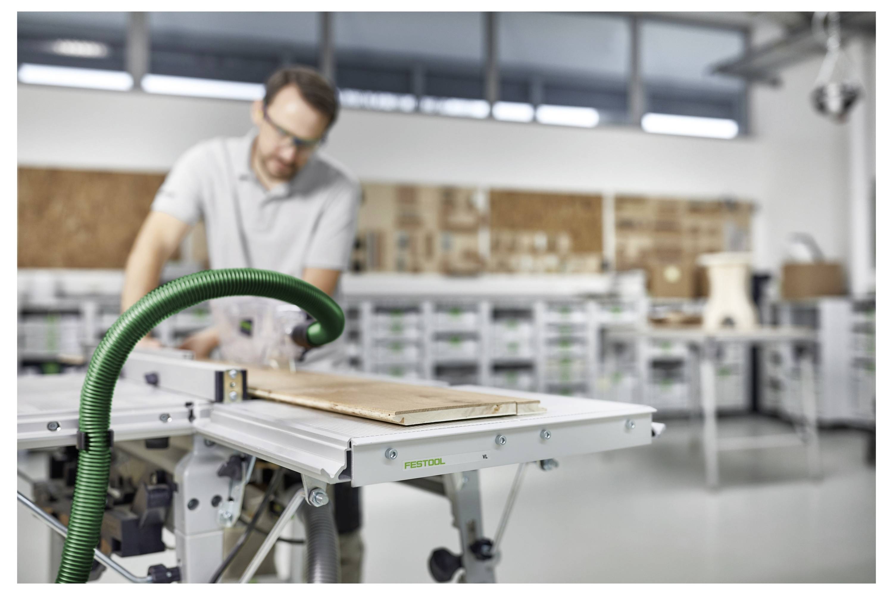 Eine Person benutzt eine Tischkreissäge mit einem grünen Staubsaugerschlauch, der angeschlossen ist, um ein Stück Holz in einer Werkstatt zu schneiden.