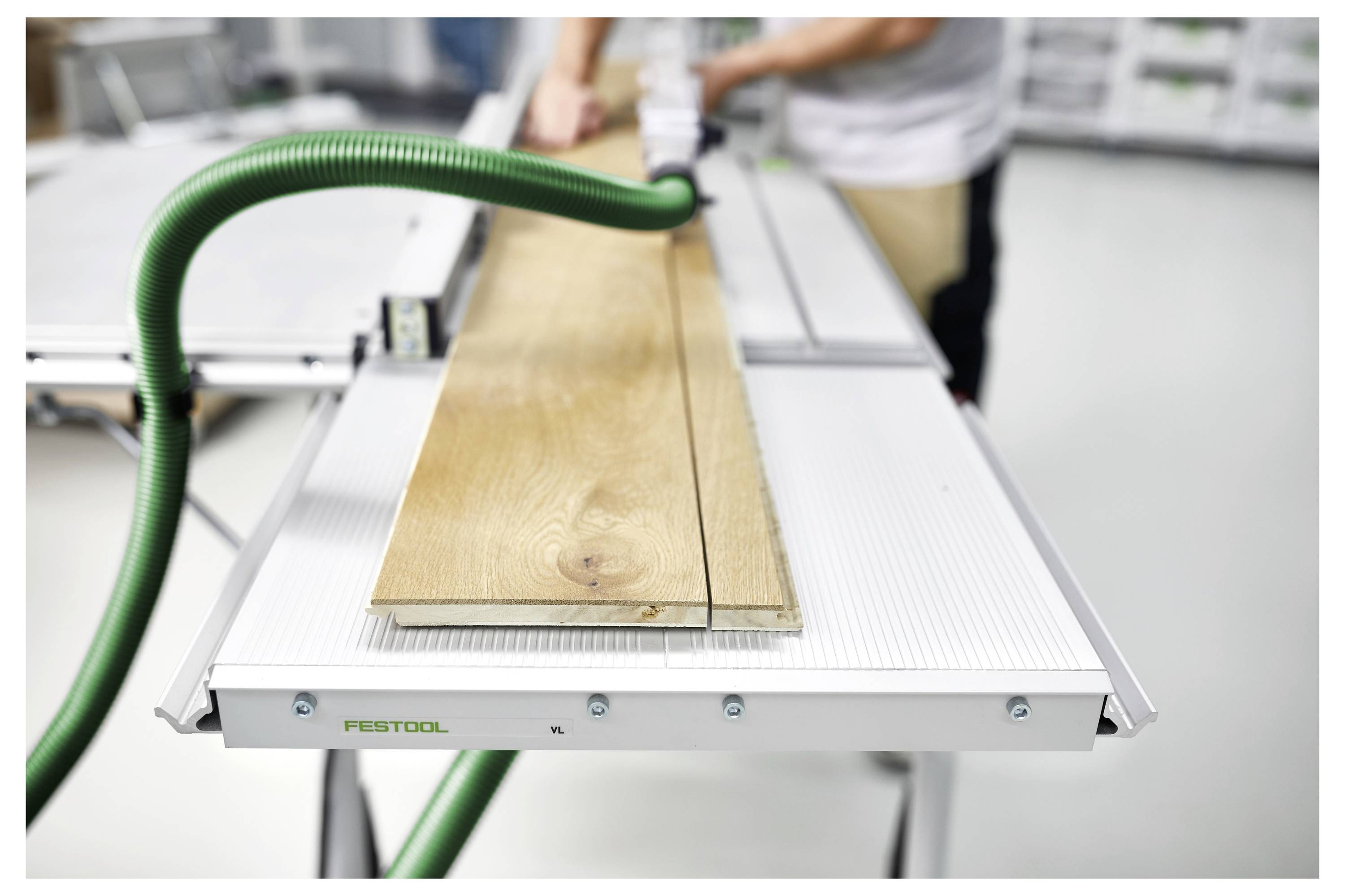 Eine Person schneidet ein Holzbrett auf einer Tischkreissäge mit einem grünen Staubabsaugschlauch zur Staubentfernung in einer Werkstatt.