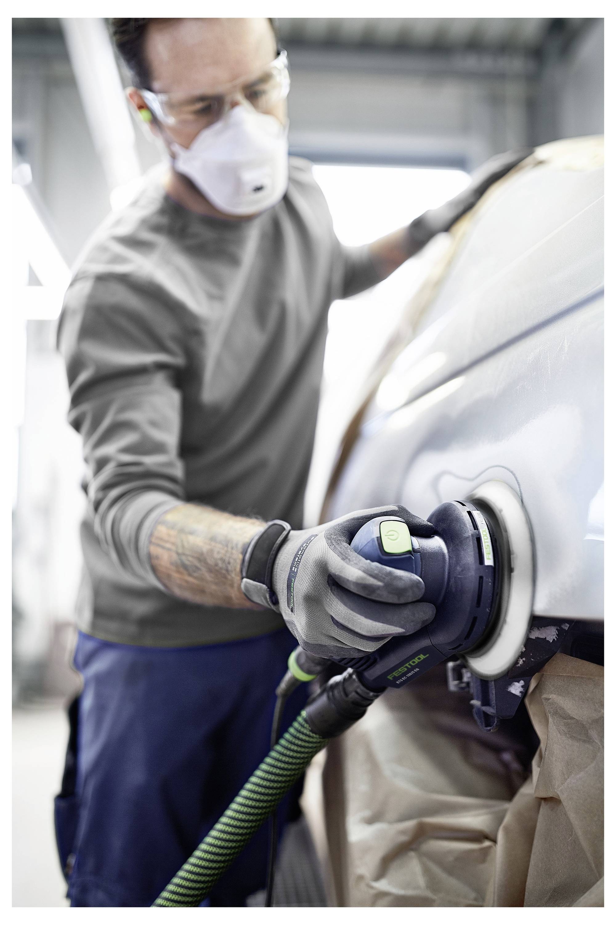 Eine Person in Schutzausrüstung poliert die Oberfläche eines Autos mit einem Elektrowerkzeug in einer Werkstatt.