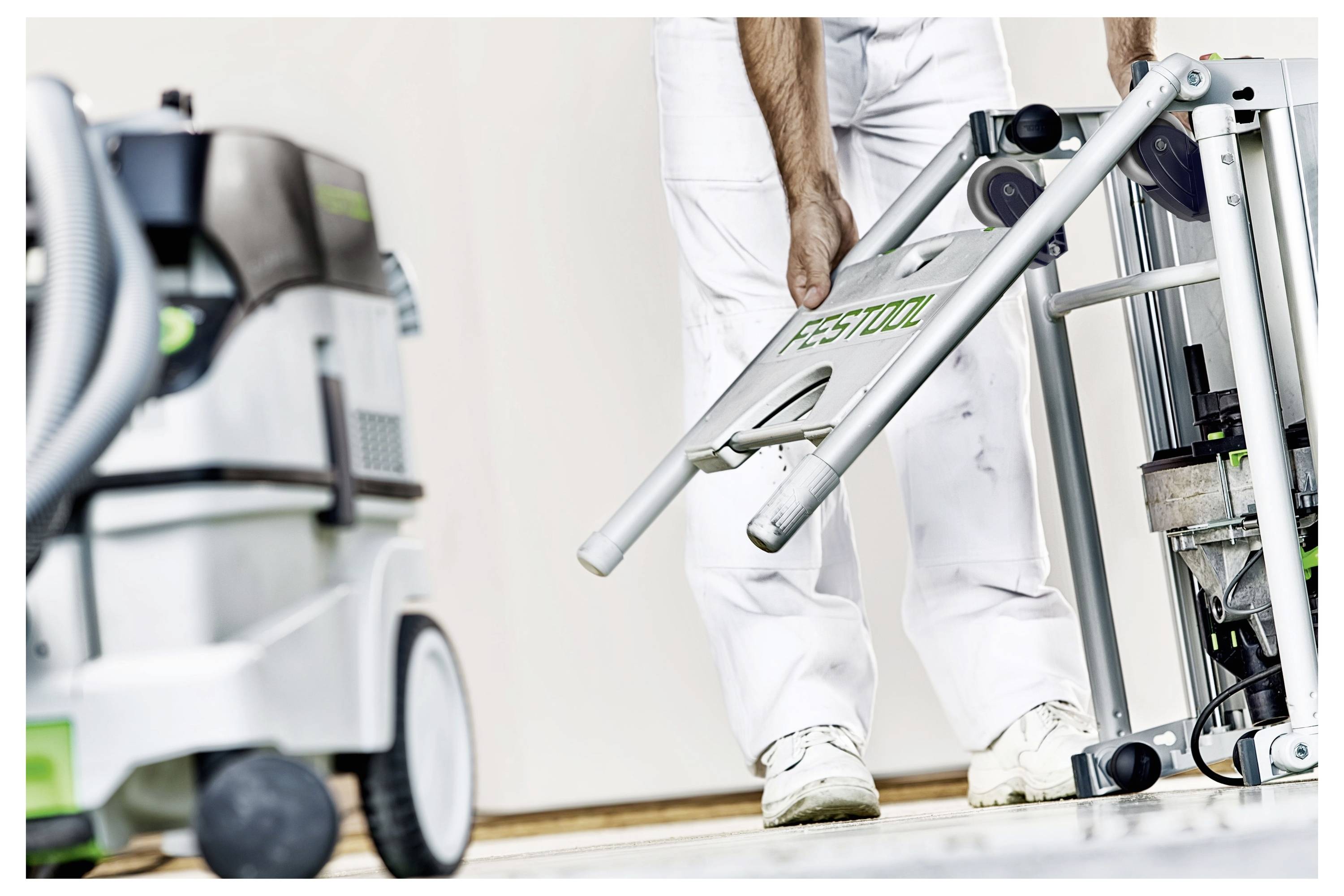 Eine Person in weißem Arbeitsoverall montiert oder justiert eine Festool-Maschine, während sich ein weiterer Festool-Staubsauger im Vordergrund befindet.