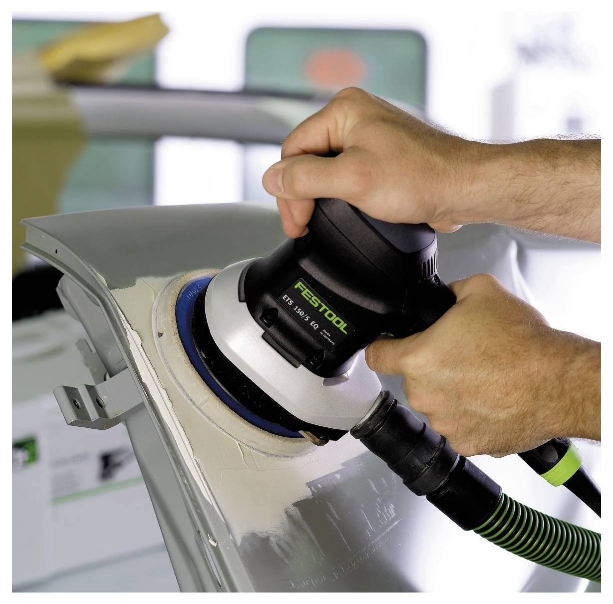 Nahaufnahme von Händen, die einen elektrischen Schleifer auf einem lackierten Autoteil verwenden, um die Oberfläche zu glätten. Das Werkzeug ist schwarz und rund mit einem grünen Schlauch.