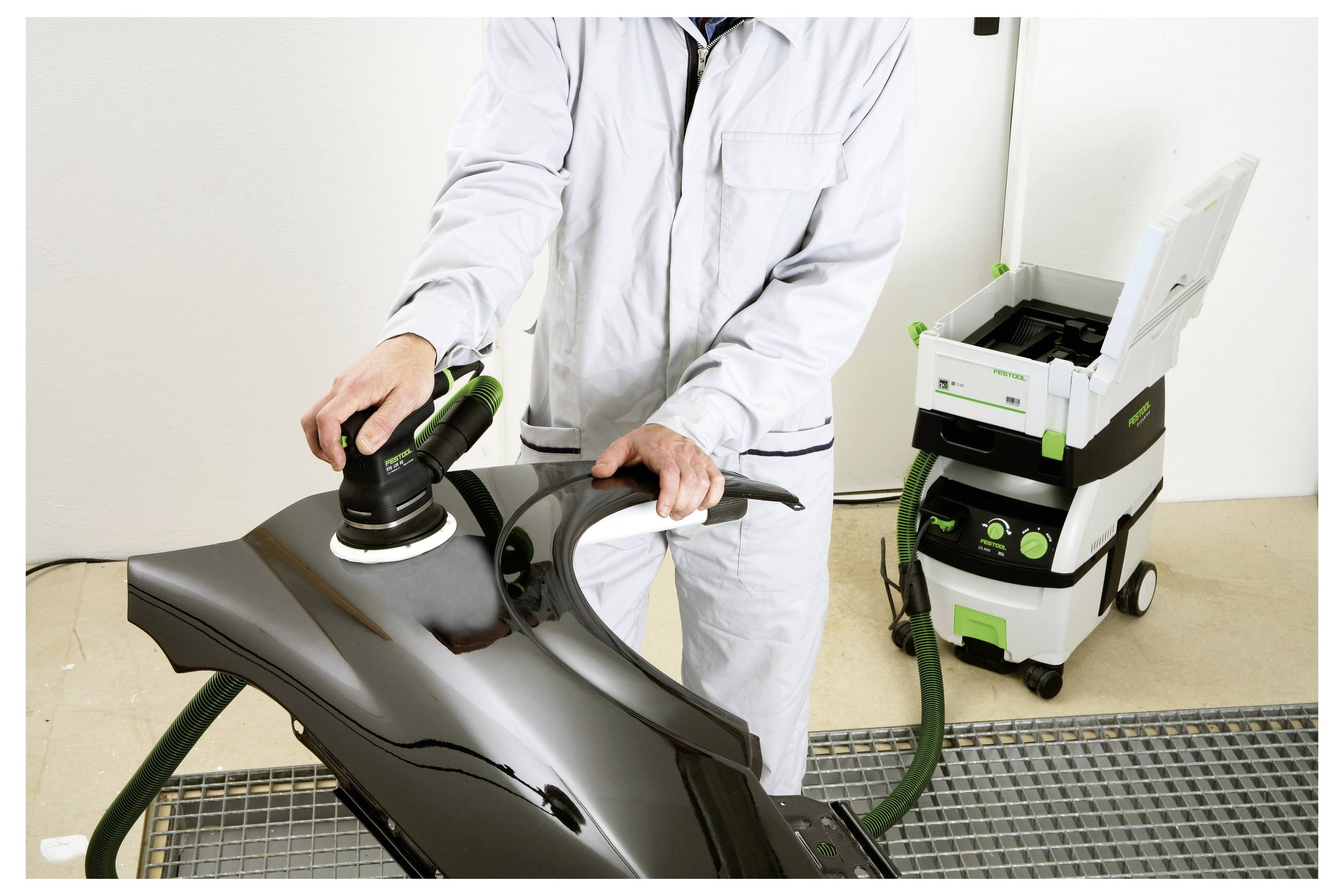 Person in a white suit using a polishing tool on a black car part connected to a vacuum system.<br><br>---<br><br>Person in einem weißen Anzug, der ein Polierwerkzeug auf einem schwarzen Autoteil verwendet, das mit einem Vakuumsystem verbunden ist.