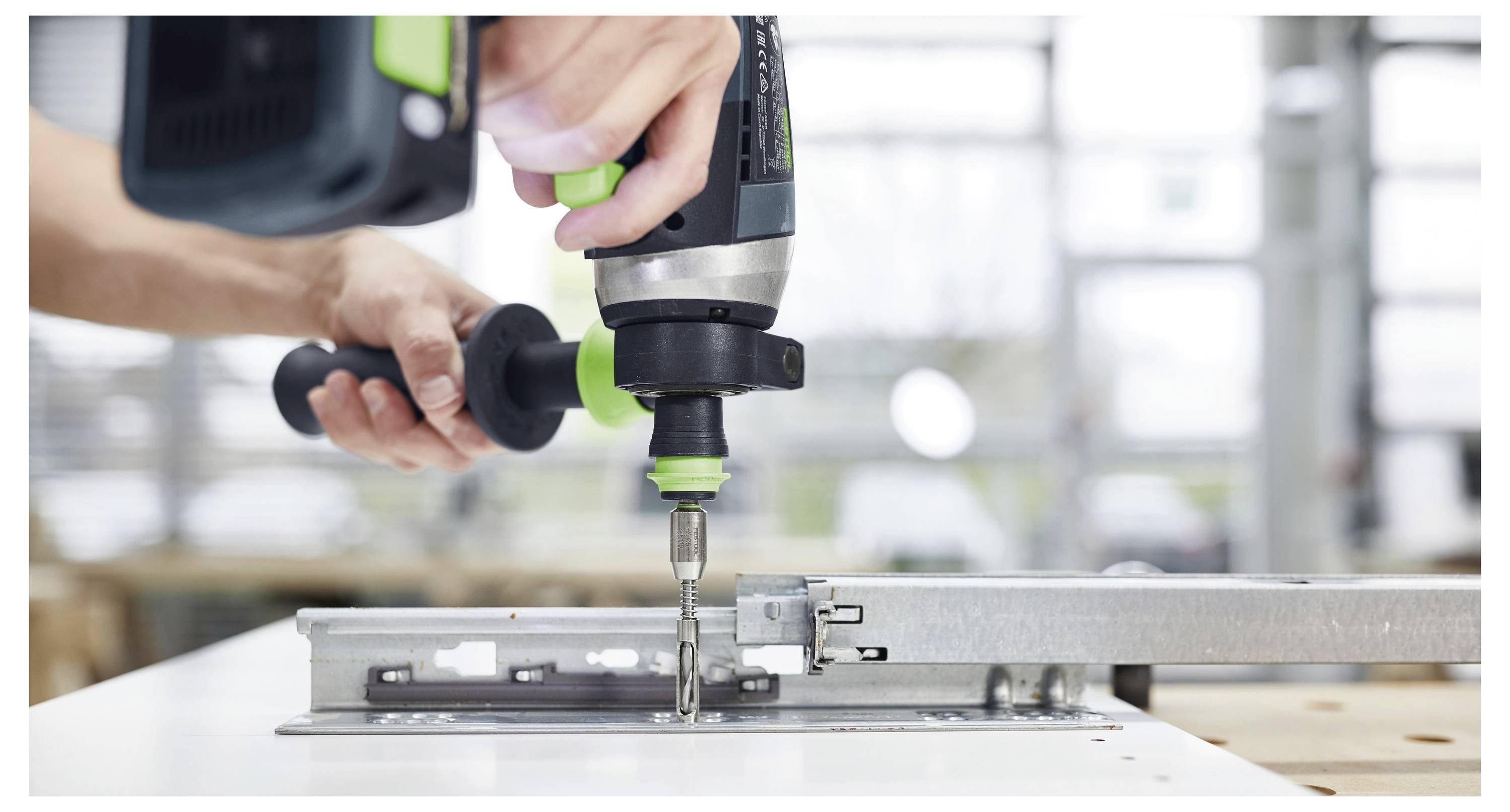 Eine Person benutzt einen Elektroschrauber, um eine Metallhalterung an einer weißen Oberfläche in einer Werkstattumgebung zu befestigen.