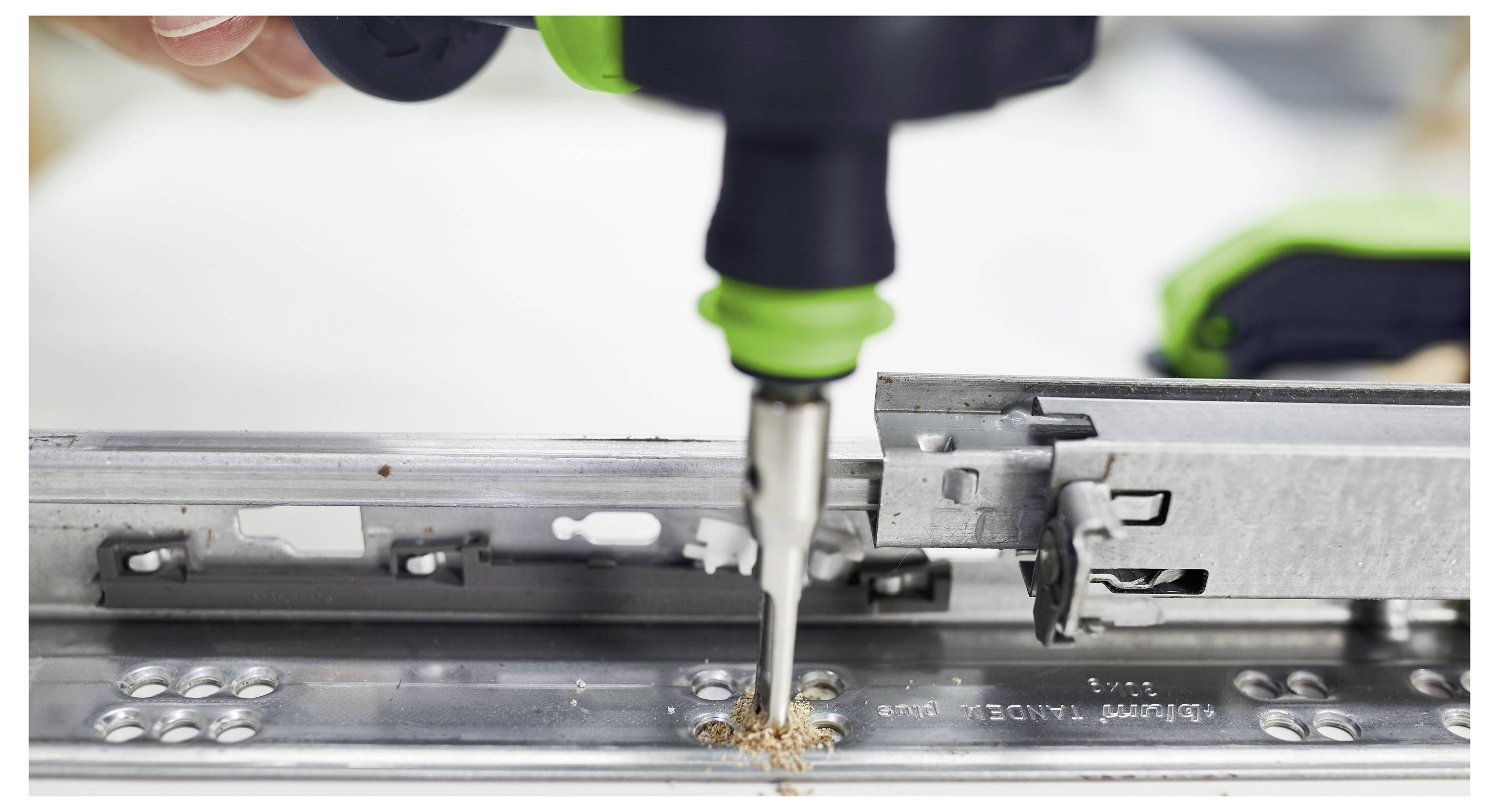 Eine Nahaufnahme eines Bohrers, der ein Loch in eine Metallschiene bohrt, mit sichtbaren Holzspänen. Der Bohrer wird von einer Hand gehalten, nah am Fokus.