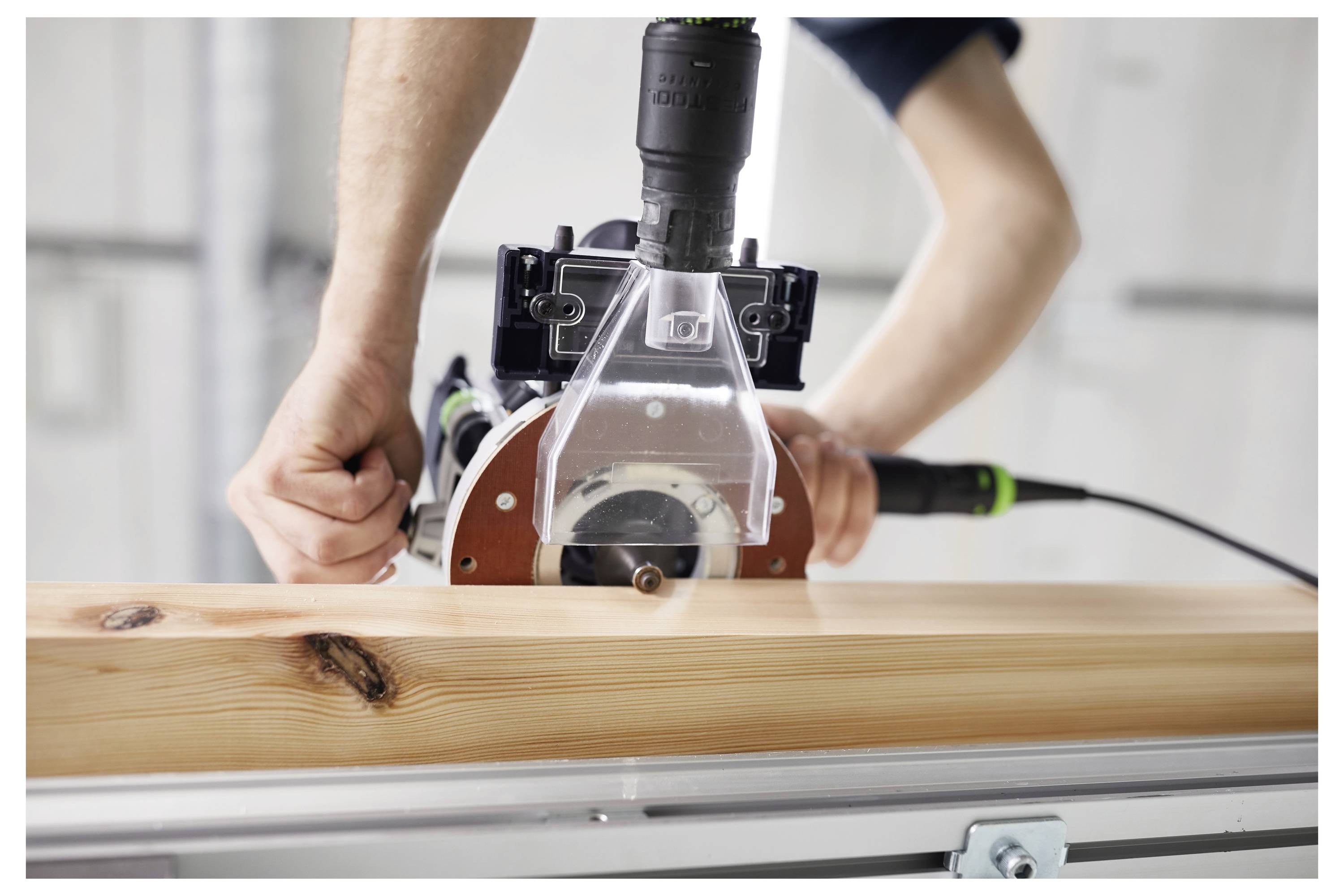 Eine Person benutzt ein Elektrowerkzeug, um ein Stück Holz zu schneiden. Das Werkzeug ist mit einer durchsichtigen Schutzvorrichtung ausgestattet. Das Holz hat ein natürliches Maserungsmuster.