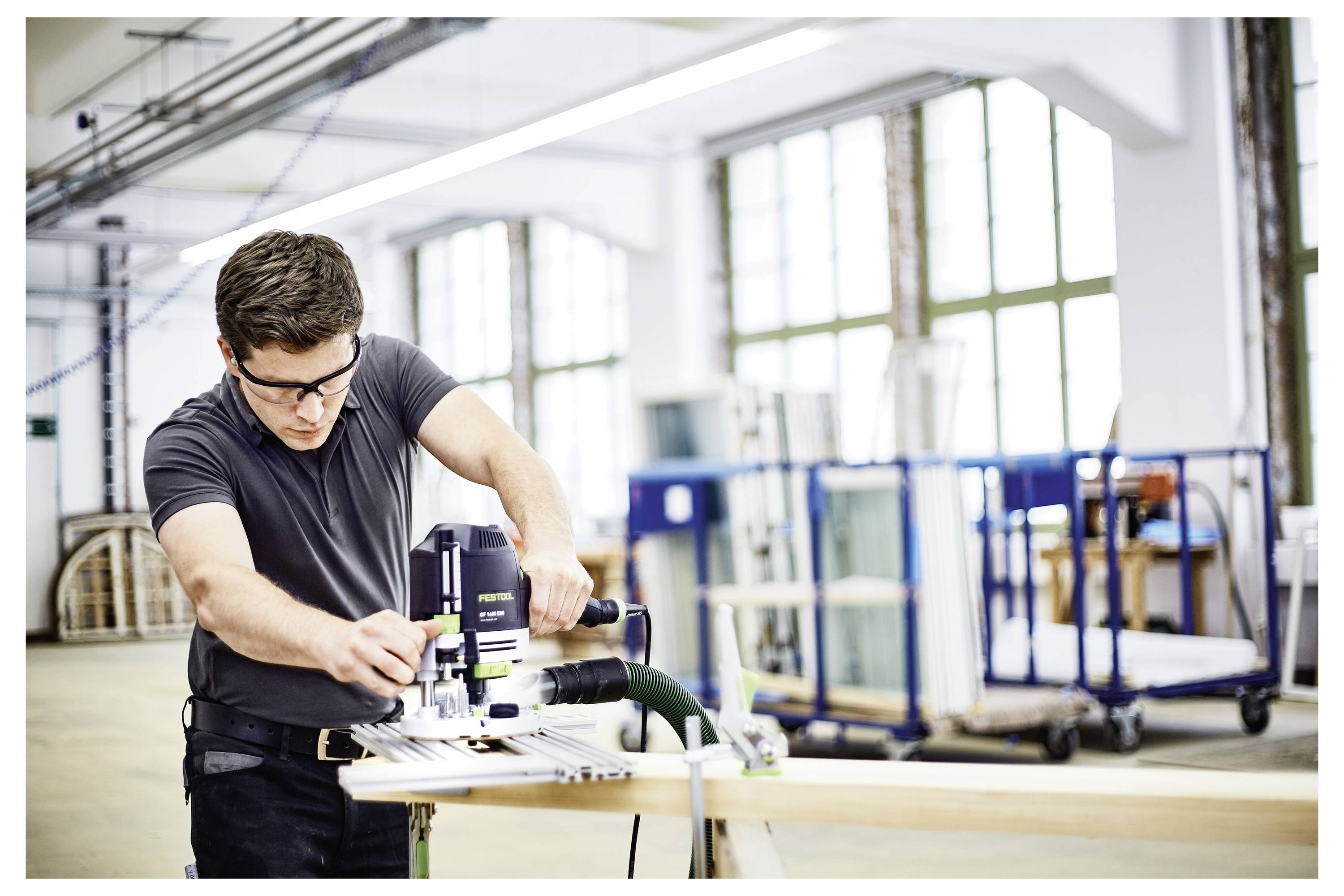 Eine Person in einer Werkstatt nutzt ein Elektrowerkzeug, um Holz zu schneiden. Der Hintergrund zeigt große Fenster und Lagerwagen.
