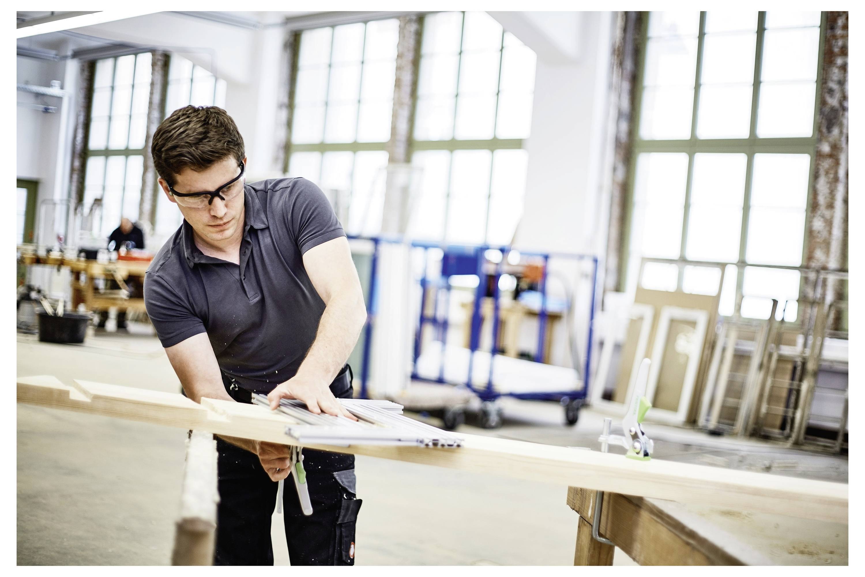 Eine Person in einer Werkstatt misst ein Holzbrett, trägt Schutzbrille und ein dunkles Hemd, konzentriert sich auf präzise Holzbearbeitungsaufgaben.