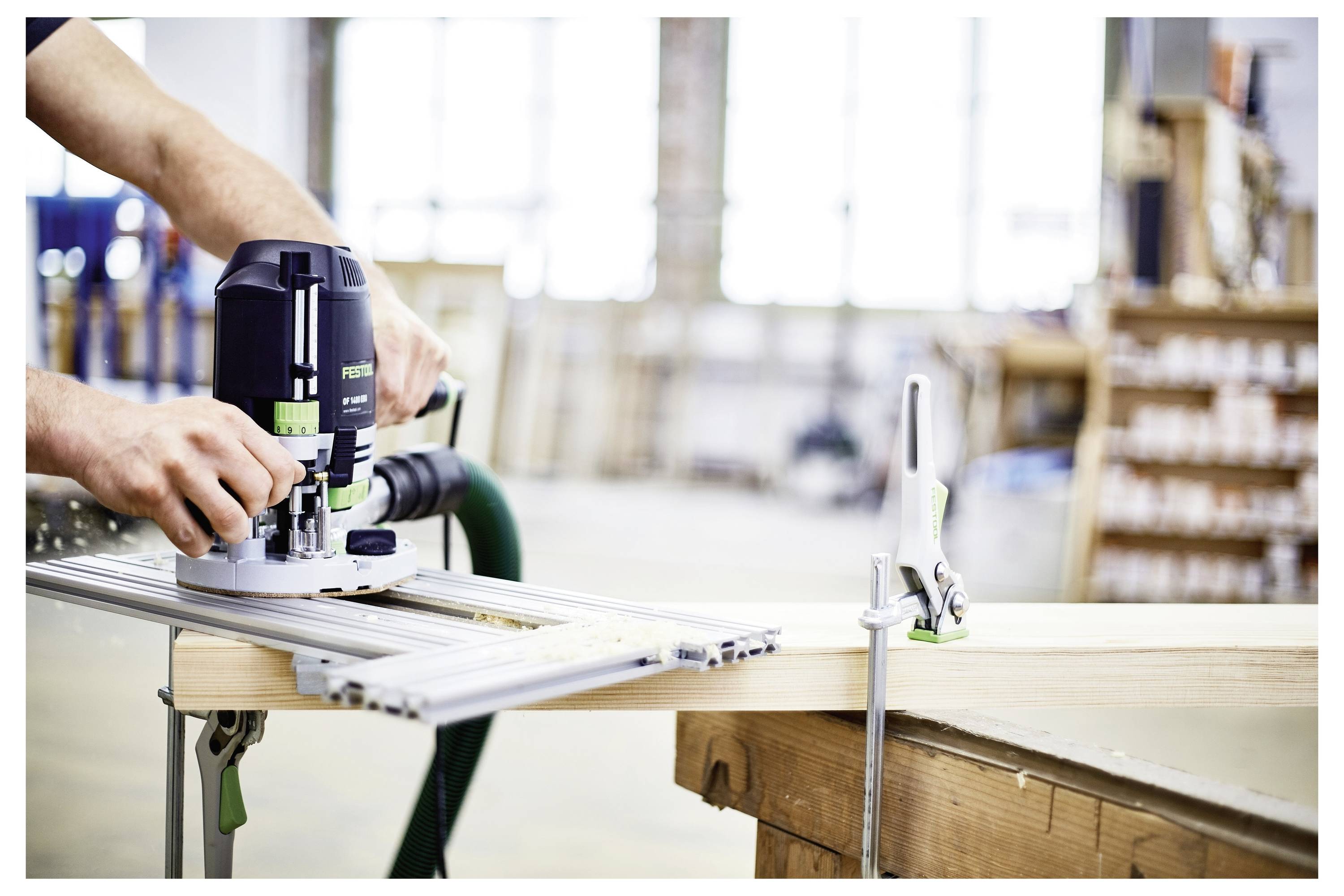 Eine Person nutzt einen elektrischen Oberfräser auf einer Holzwerkbank in einer Werkstatt, wobei sie sich auf präzise Holzbearbeitung mit Werkzeugen und Spannzwingen in der Nähe konzentriert.