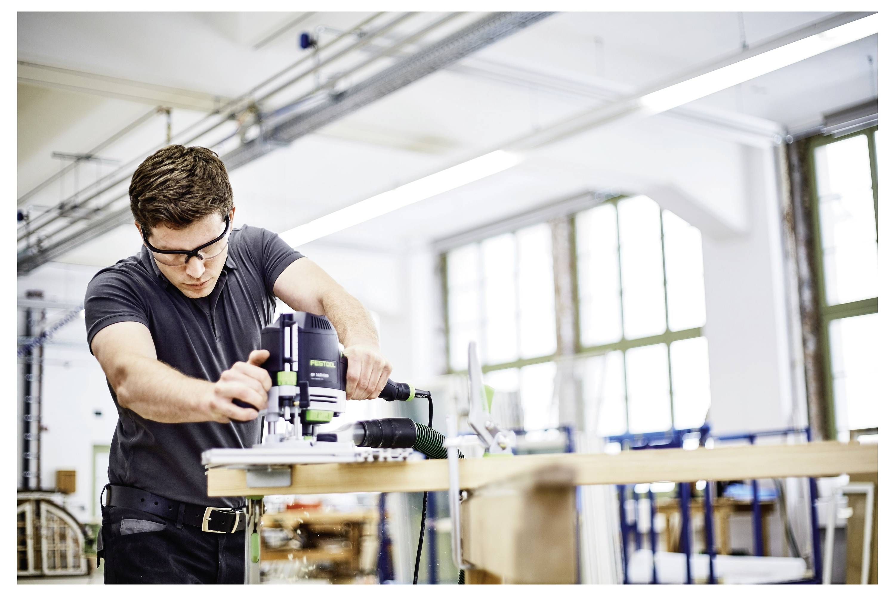 Ein Mann, der eine Schutzbrille trägt, verwendet ein Elektrowerkzeug auf einem Holzbrett in einer Werkstatt und konzentriert sich intensiv auf seine Aufgabe inmitten einer Industrieumgebung.