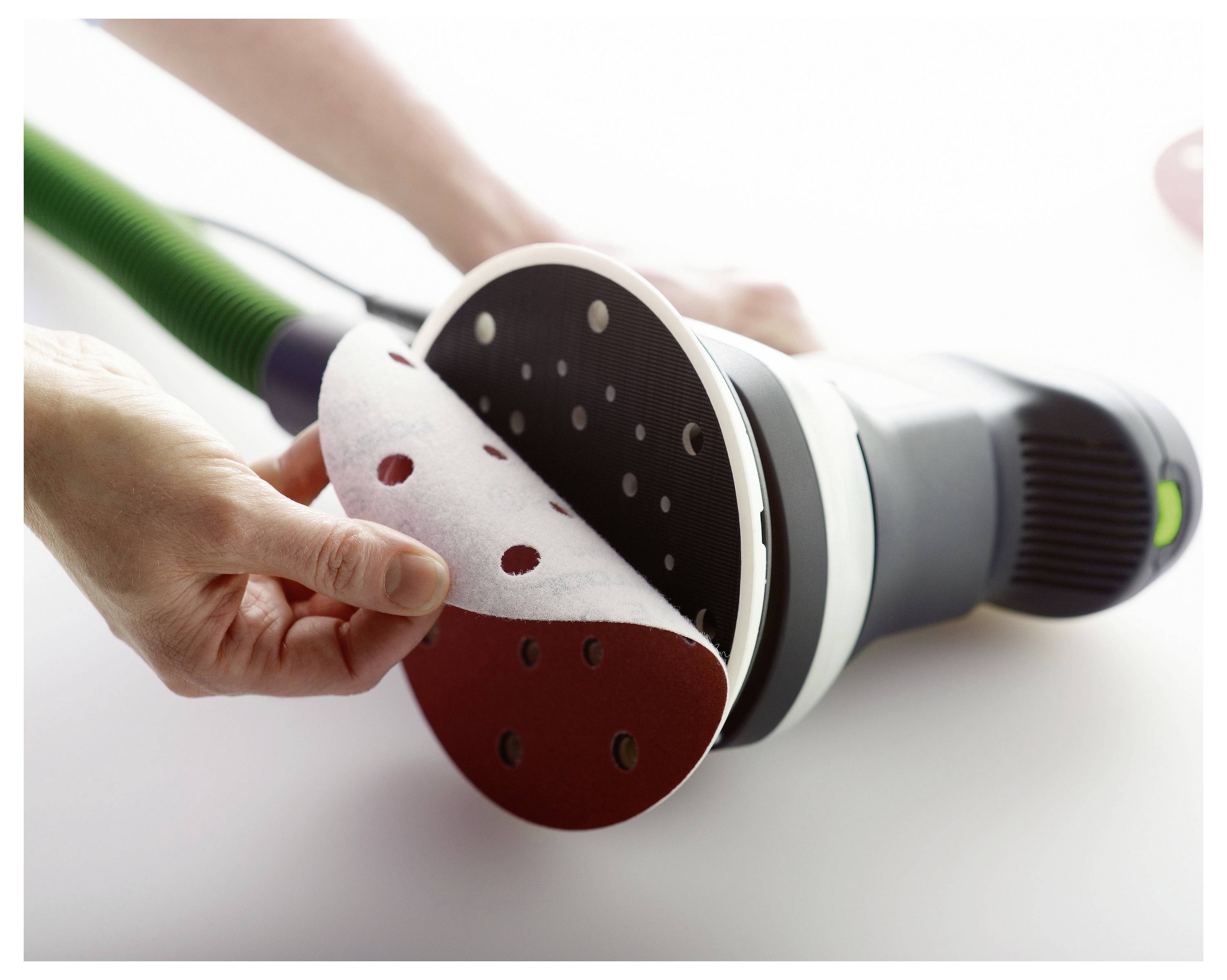 Eine Nahaufnahme von Händen, die Schleifpapier an einem Elektroschleiffer wechseln, mit Darstellung der Ausrichtung von Löchern zur Staubabsaugung auf einer weißen Oberfläche.