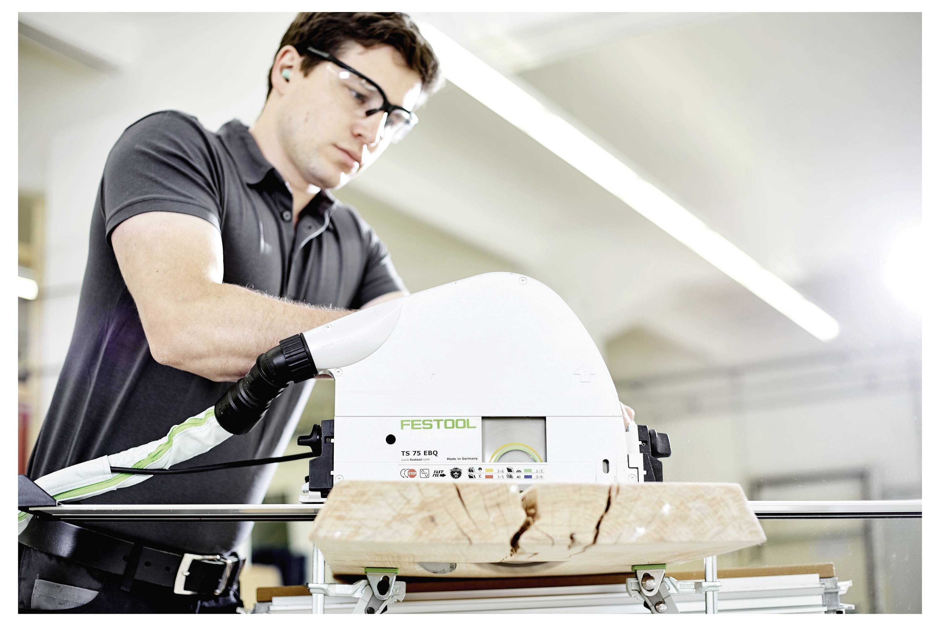 Eine Person mit Schutzbrille bedient eine Festool TS 55 RQ Kreissäge und sägt ein Stück Holz auf einer Werkbank in einer Werkstatt zu.