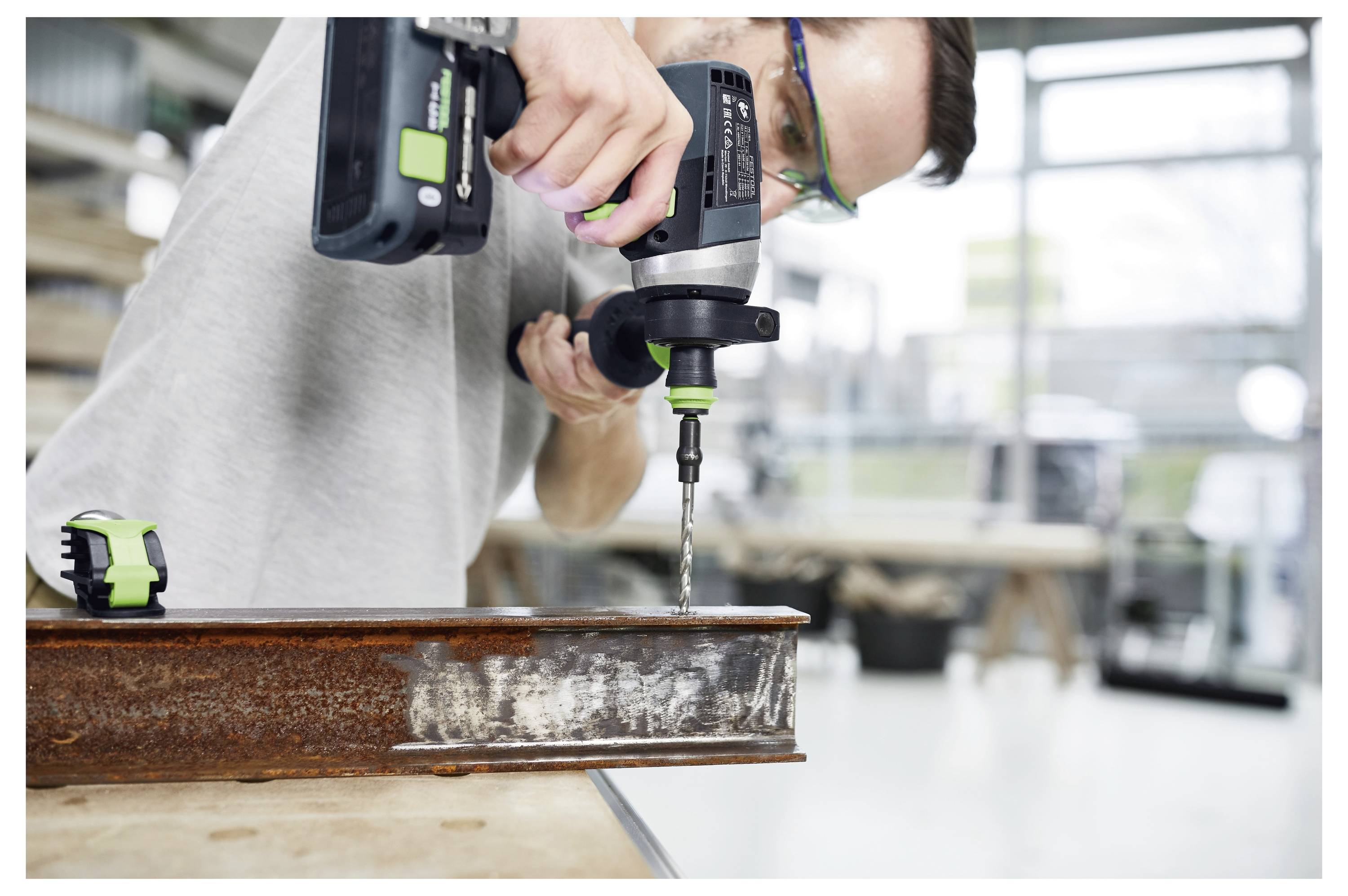 Eine Person benutzt einen kabellosen Bohrer mit einem grünen Bohrbit, um in einen Metallbalken in einer Werkstatt mit verschiedenen Werkzeugen im Hintergrund zu bohren.