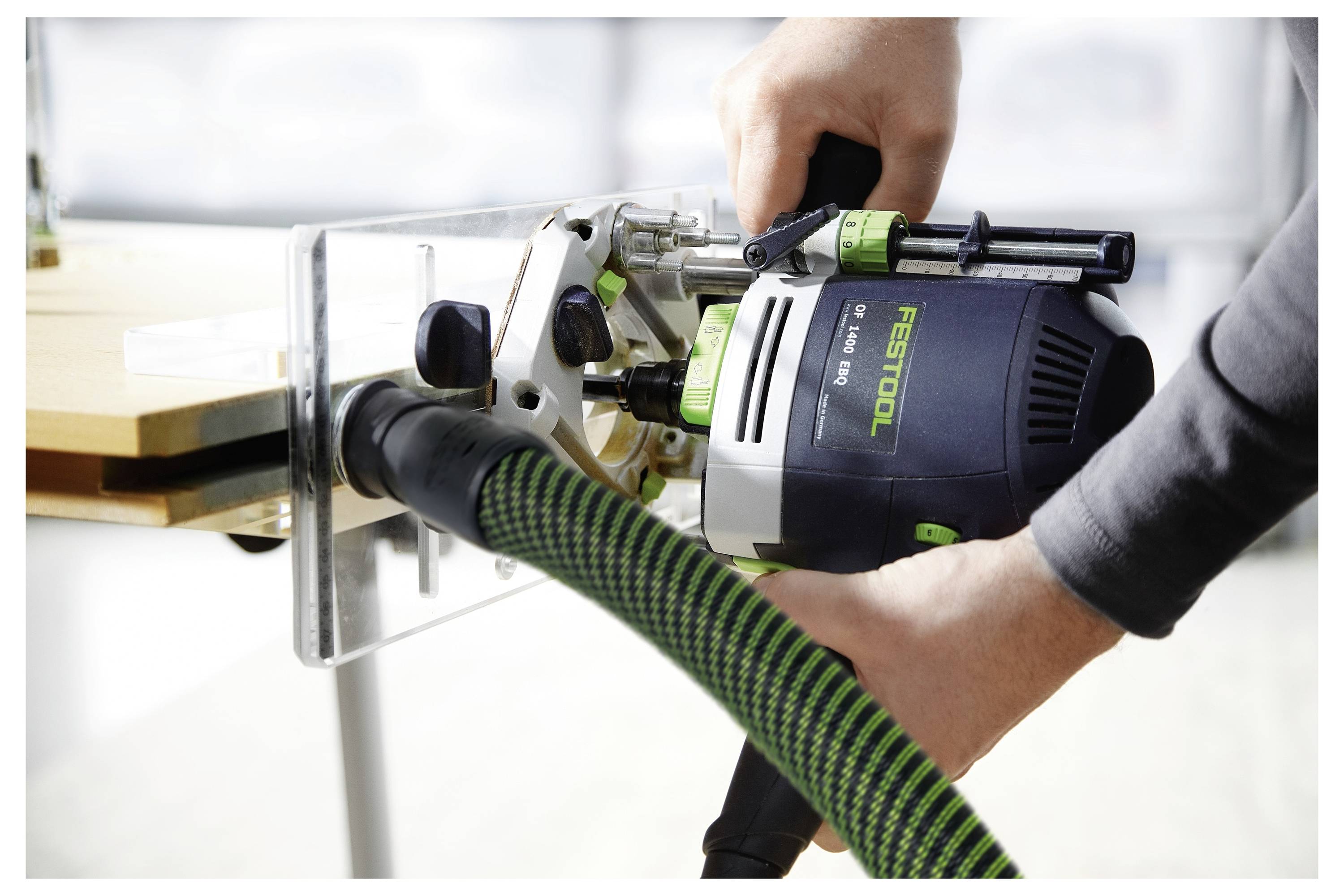 Eine Person, die einen Festool-Fräser auf einer Holzoberfläche benutzt, verbindet ihn mit einem Staubabsaugsystem mittels eines grün-schwarzen Schlauchs.