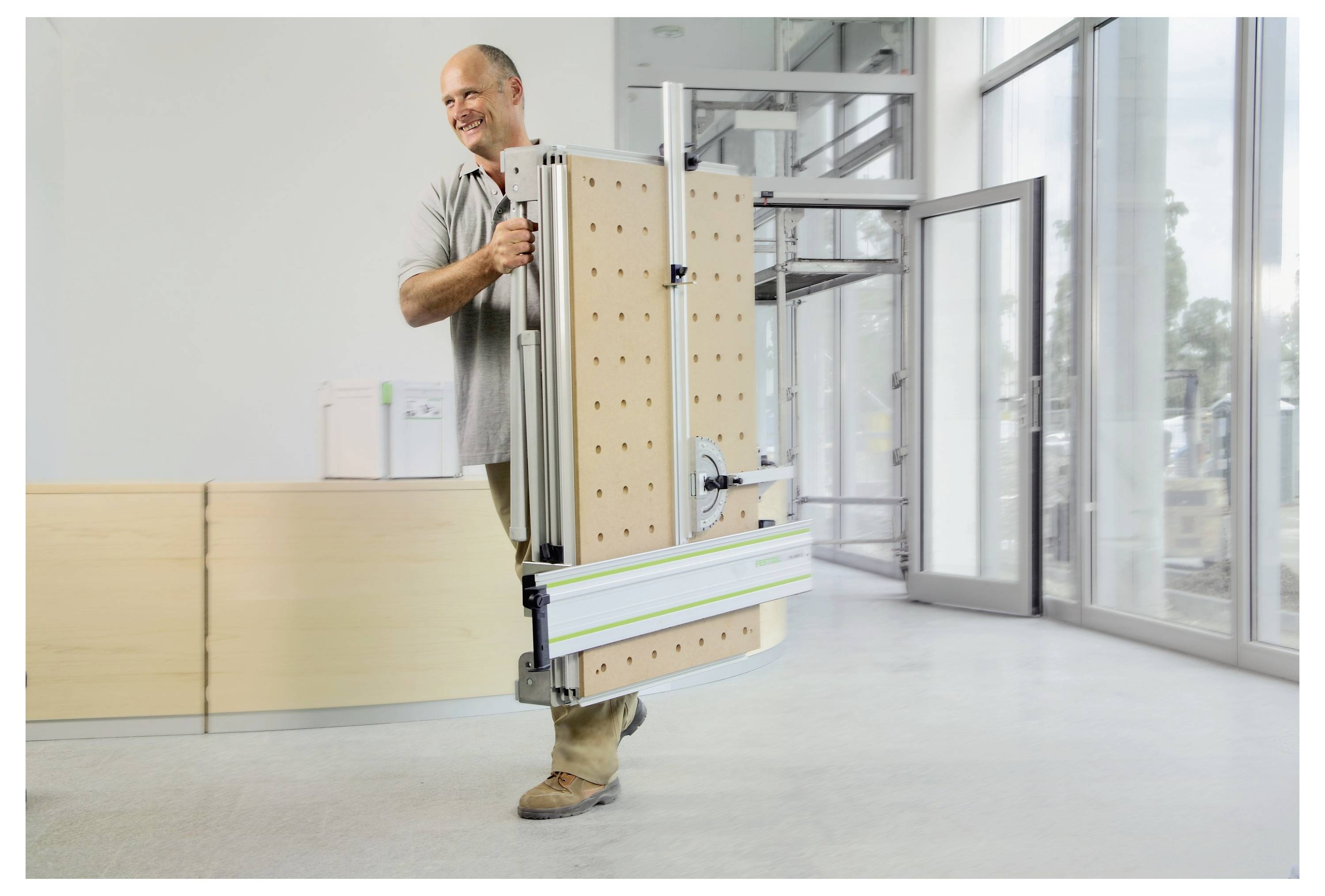 Eine Person, die lächelt, während sie eine zusammengeklappte, tragbare Werkbank mit Lochrasterseiten in einem hellen, modernen Büroraum mit Glasfenstern trägt.