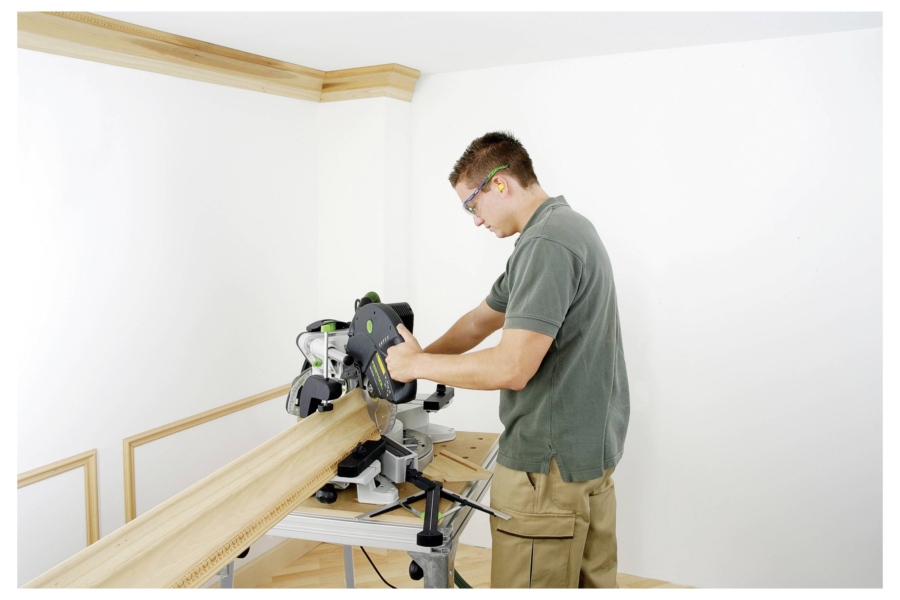 Eine Person, die in einer Werkstatt mit einer Gehrungssäge Holz schneidet, trägt eine Schutzbrille und Gehörschutz und steht neben einer Eckwand.