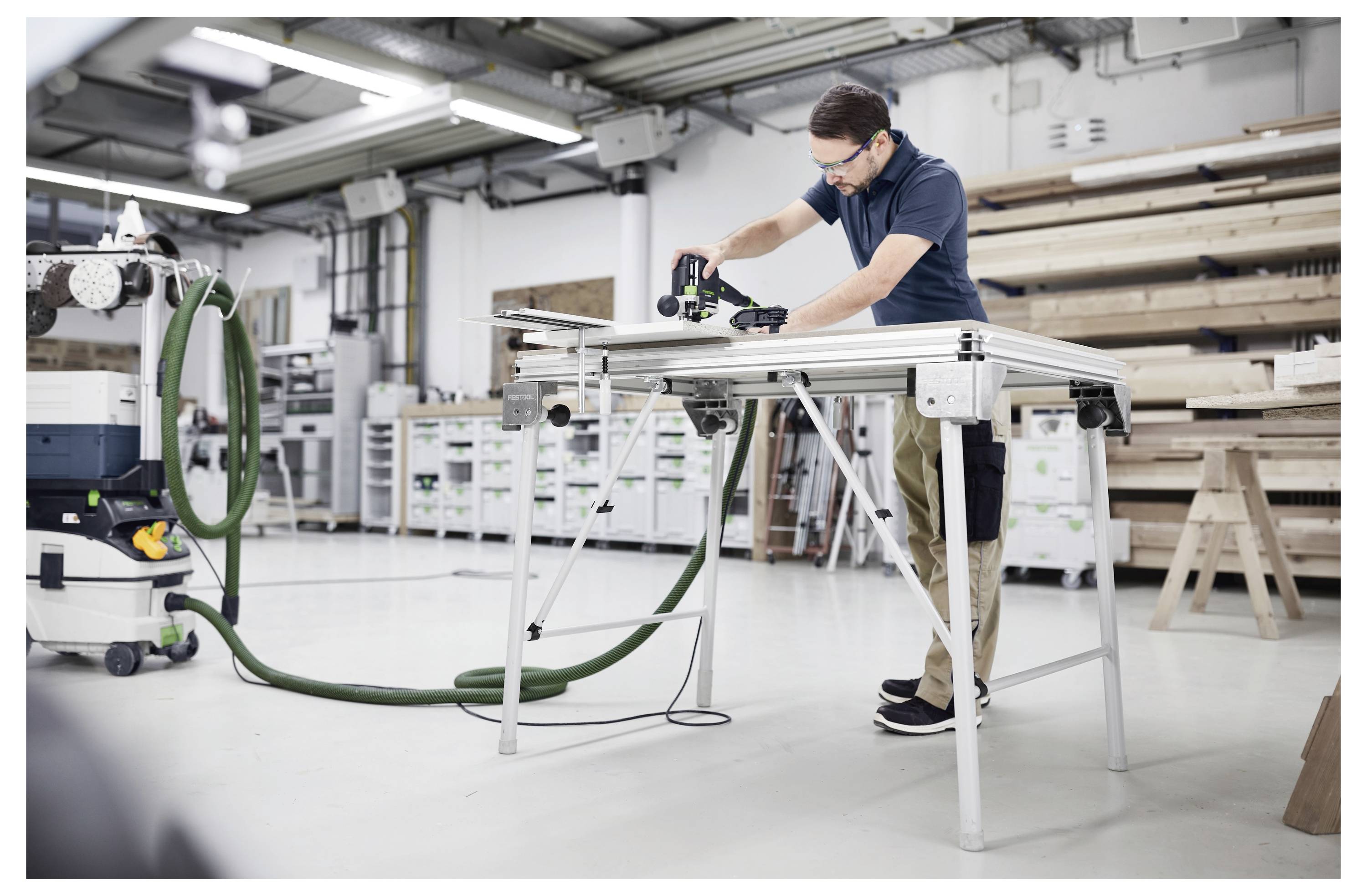 Eine Person benutzt ein Elektrowerkzeug, um Holz auf einer Werkbank in einer Werkstatt zu schneiden, die mit verschiedenen Werkzeugen und Holzmaterialien gefüllt ist.