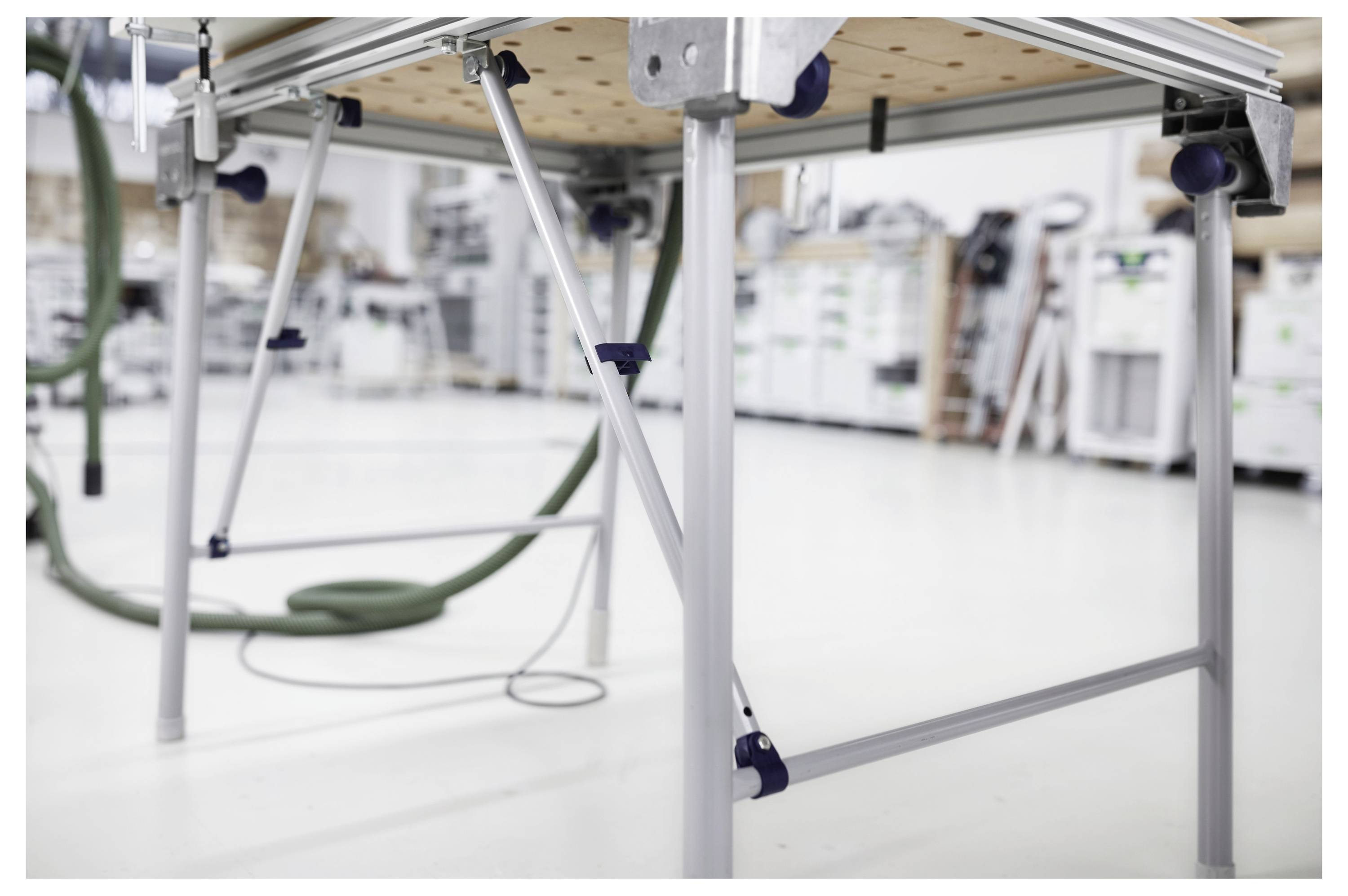 Unteransicht eines Metalltischs mit verstellbaren Beinen in einer Werkstatt. Regale und Ausrüstung sind im Hintergrund verschwommen.