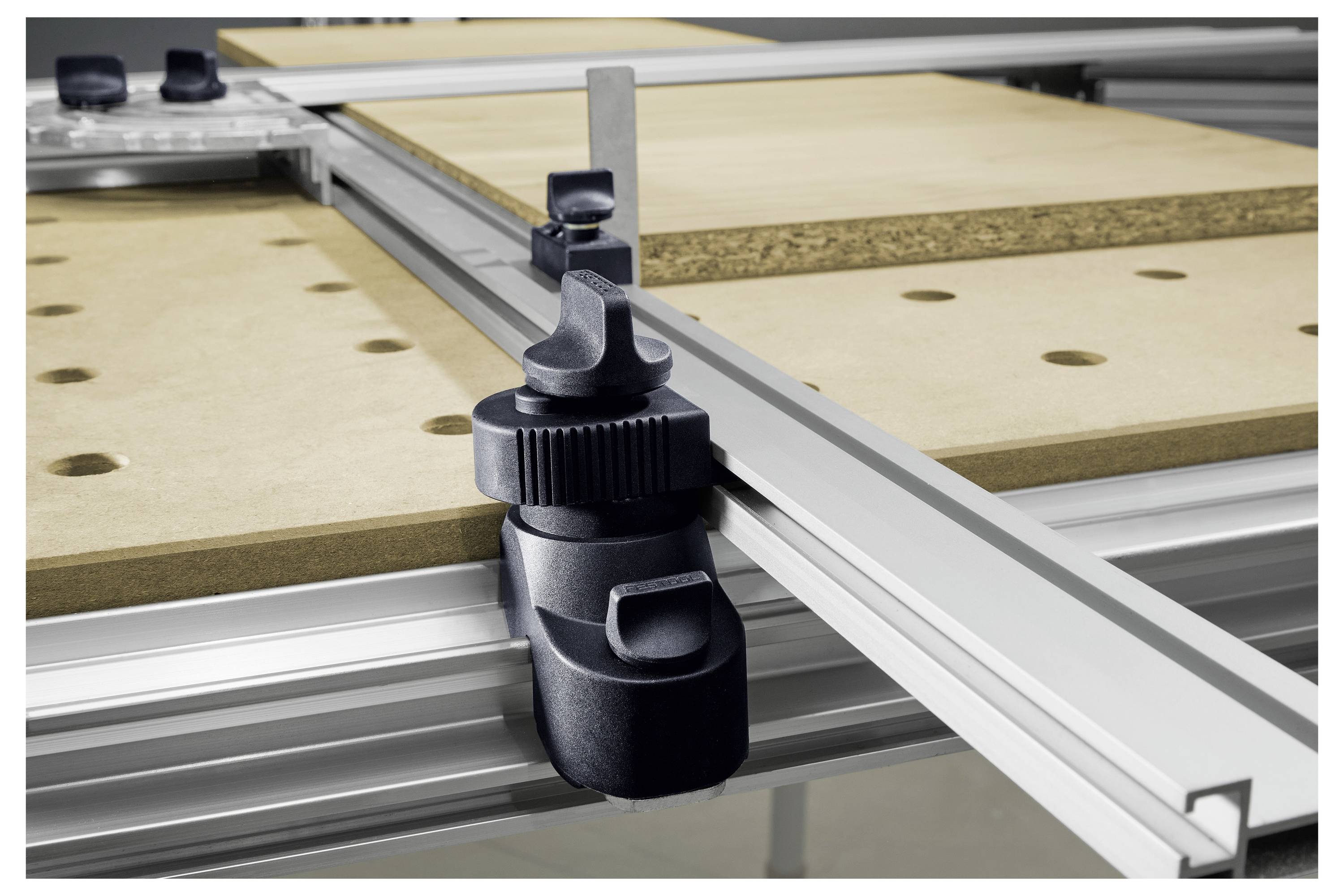 Eine Nahaufnahme einer Metallzwinge, die eine Holzplatte auf einer Werkbank sichert, mit Linealen und Führungsschienen im Hintergrund, was eine genaue Messeinrichtung anzeigt.