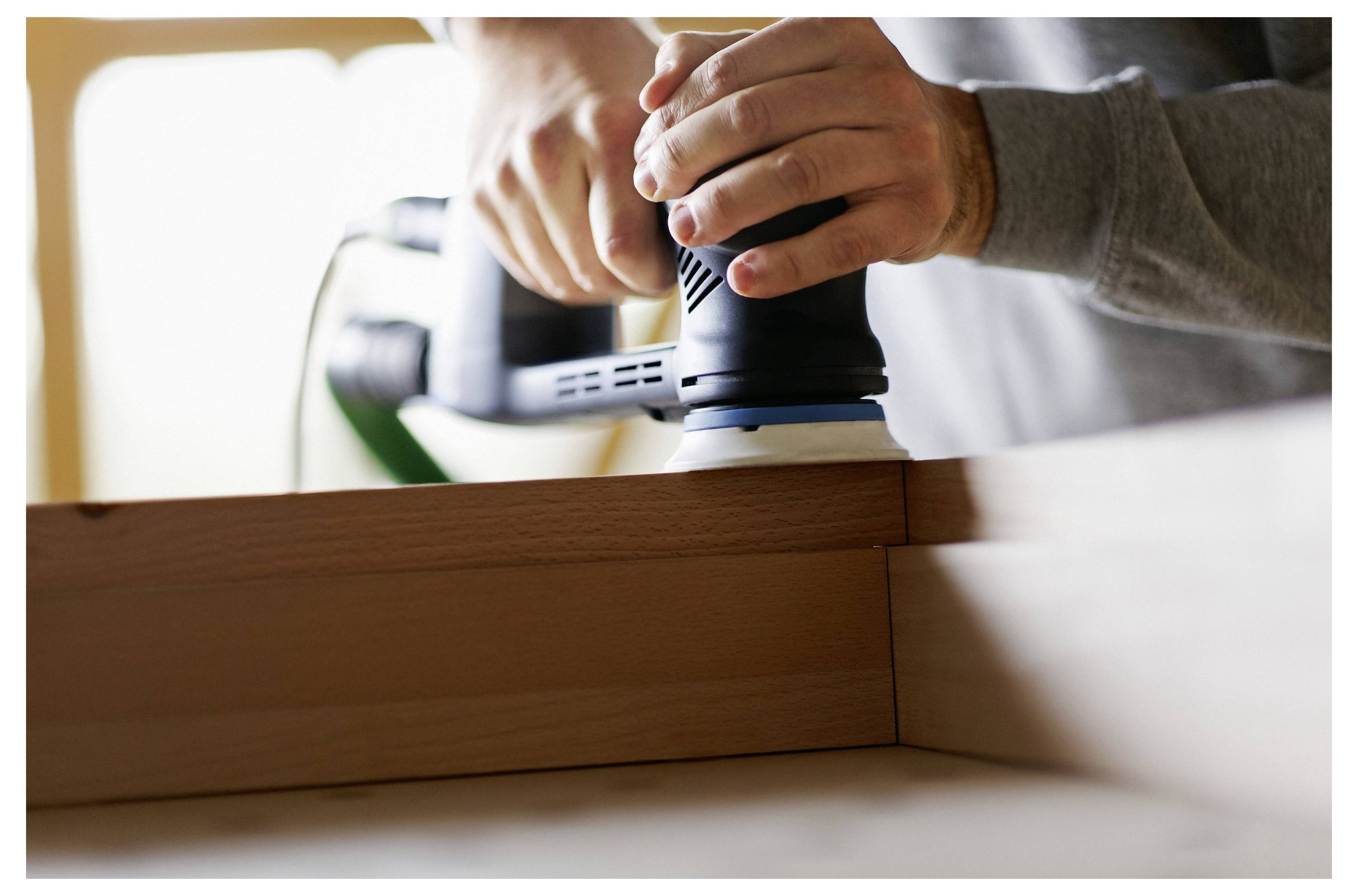 Eine Person trägt Handschuhe und schleift eine Autofläche mit einem elektrischen Schleifer und konzentriert sich darauf, die Außenseite des Fahrzeugs zum Nachschleifen zu glätten.