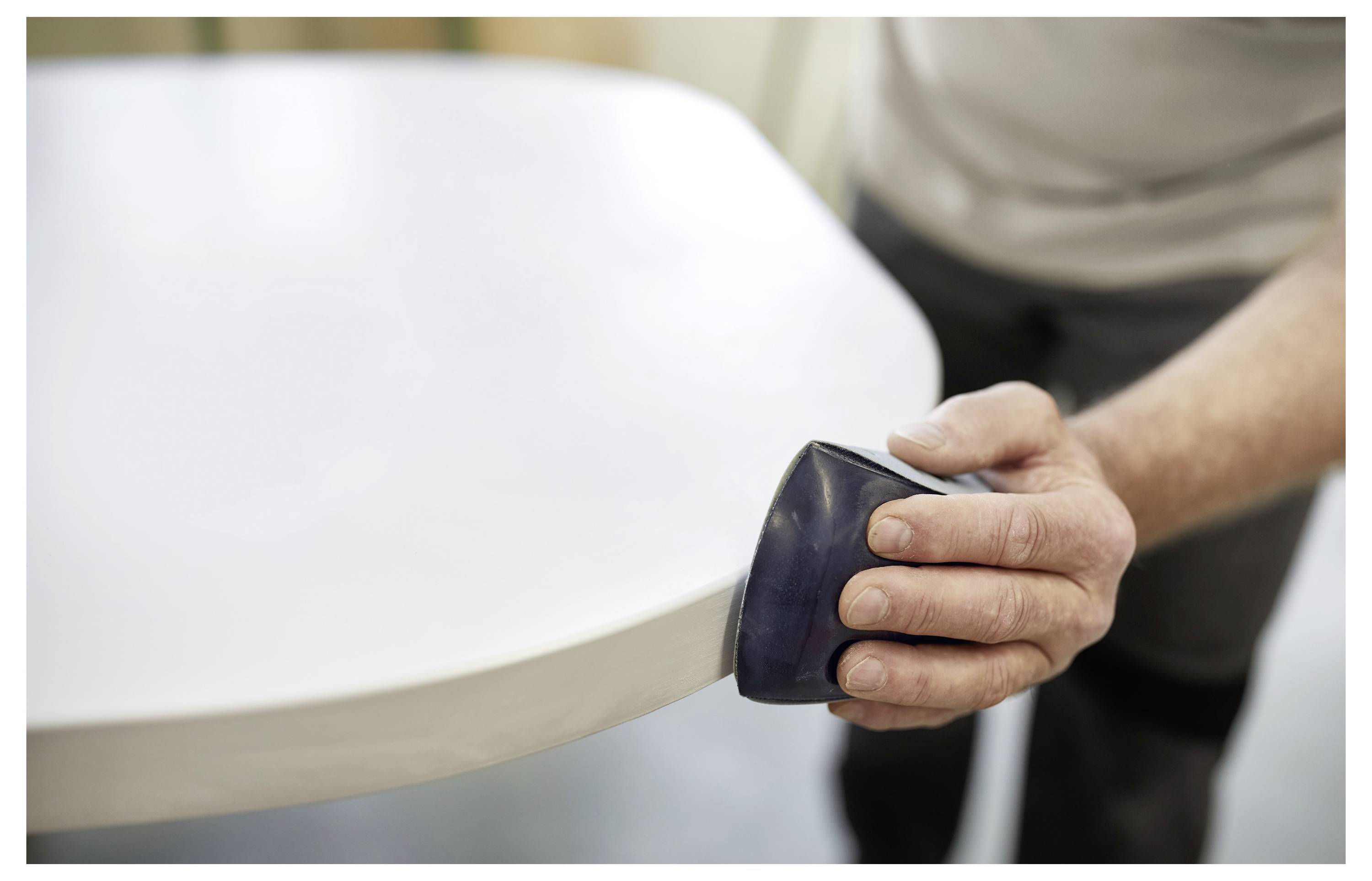 Eine Person glättet die Kante eines weißen Surfbretts mit einem Schleifwerkzeug und konzentriert sich auf präzise Handwerkskunst.