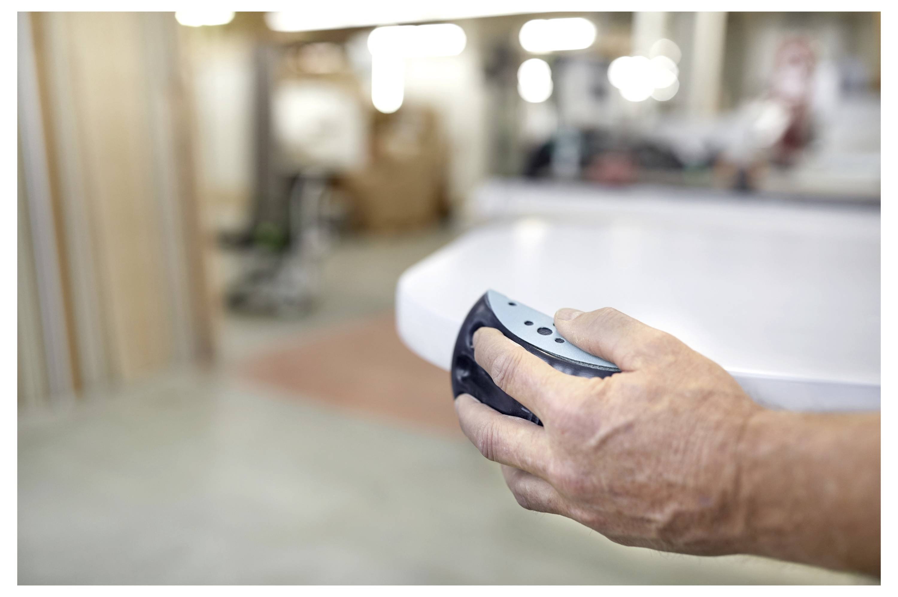 Eine Person verwendet einen blau-schwarzen tragbaren Schleifer, um die Kante eines weißen Objekts, wahrscheinlich Möbel, in einer Werkstattumgebung zu glätten.