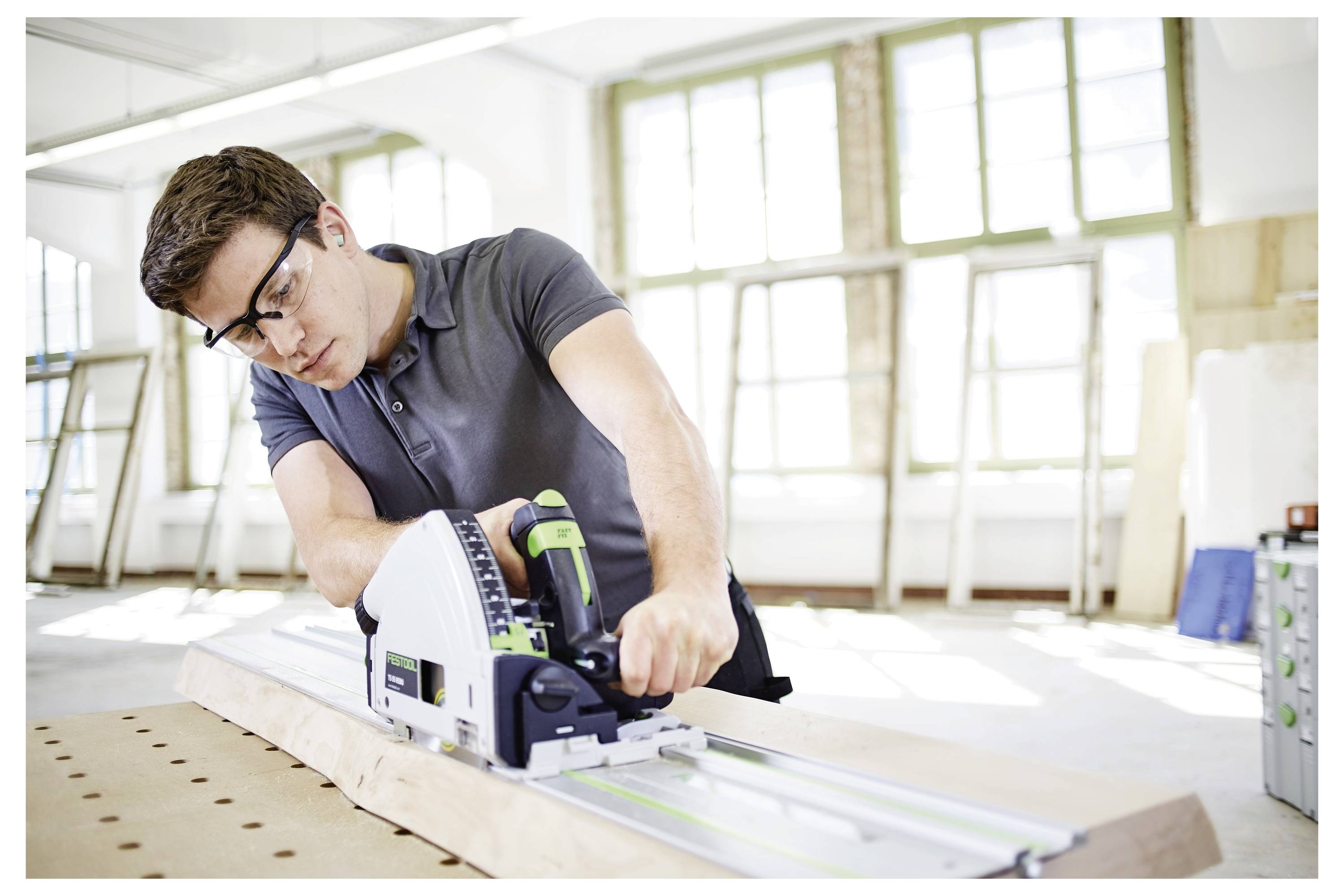 Mann, der mit einer Kreissäge Holz in einer von natürlichem Licht erfüllten Werkstatt schneidet. Er trägt eine Schutzbrille und konzentriert sich auf seine Arbeit.