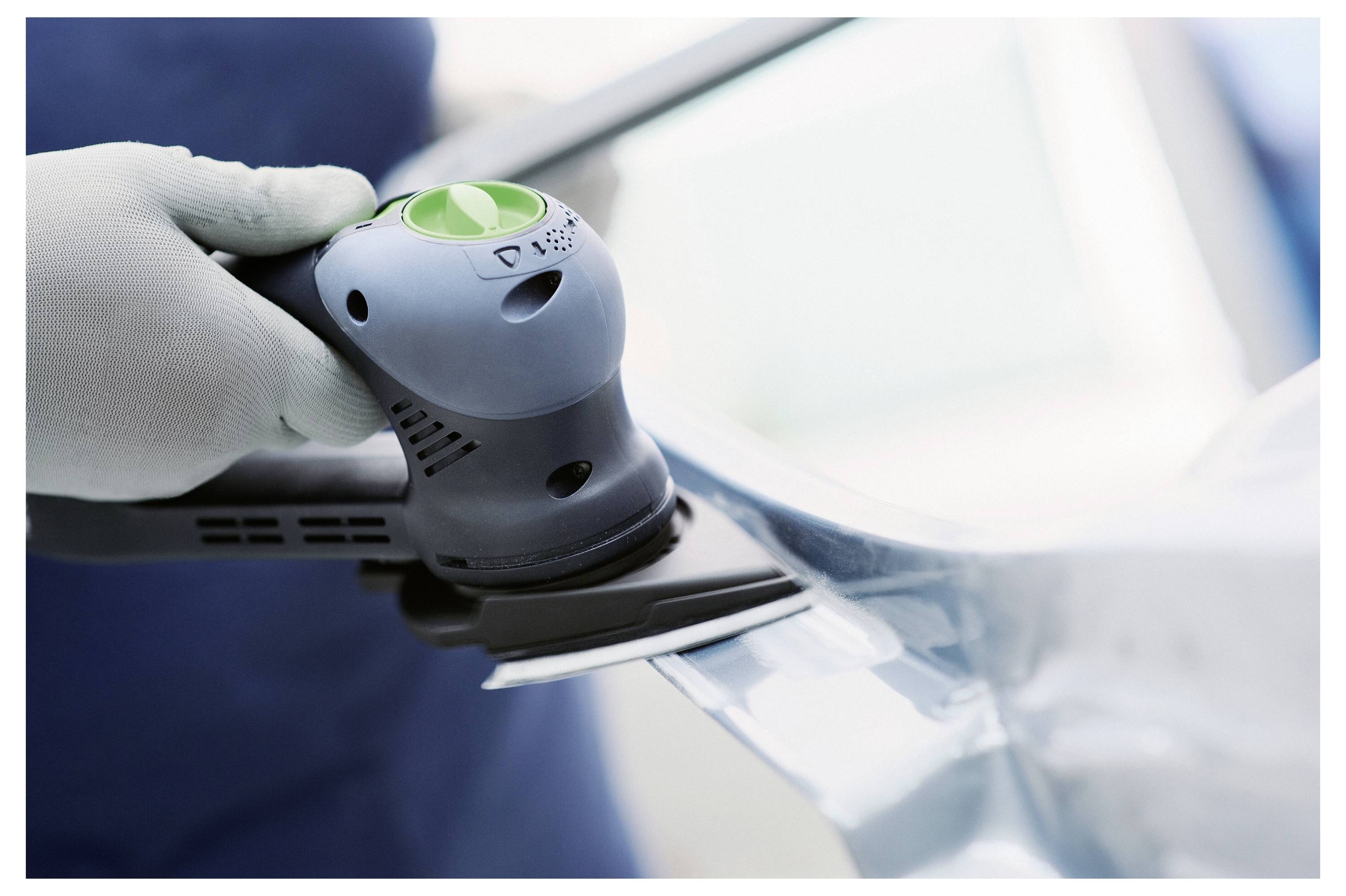 Eine Person benutzt einen tragbaren elektrischen Schleiffer auf einer glatten metallischen Oberfläche, trägt weiße Handschuhe zum Schutz und arbeitet in einer Werkstattumgebung.