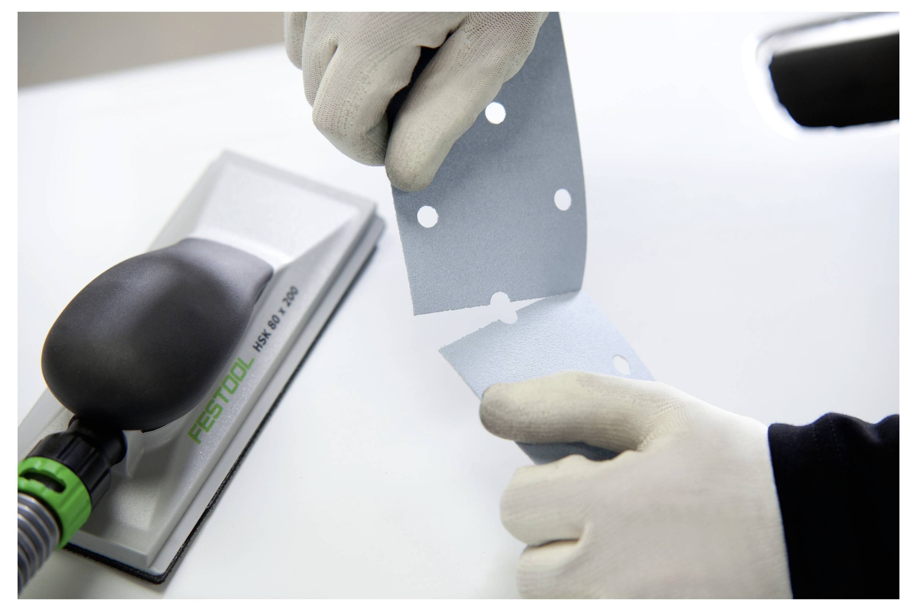 Eine Person mit Handschuhen reißt ein blaues Schleifpapierblatt mit vorgestanzten Löchern neben einem Festool-Schleifsystem auf einer weißen Oberfläche.
