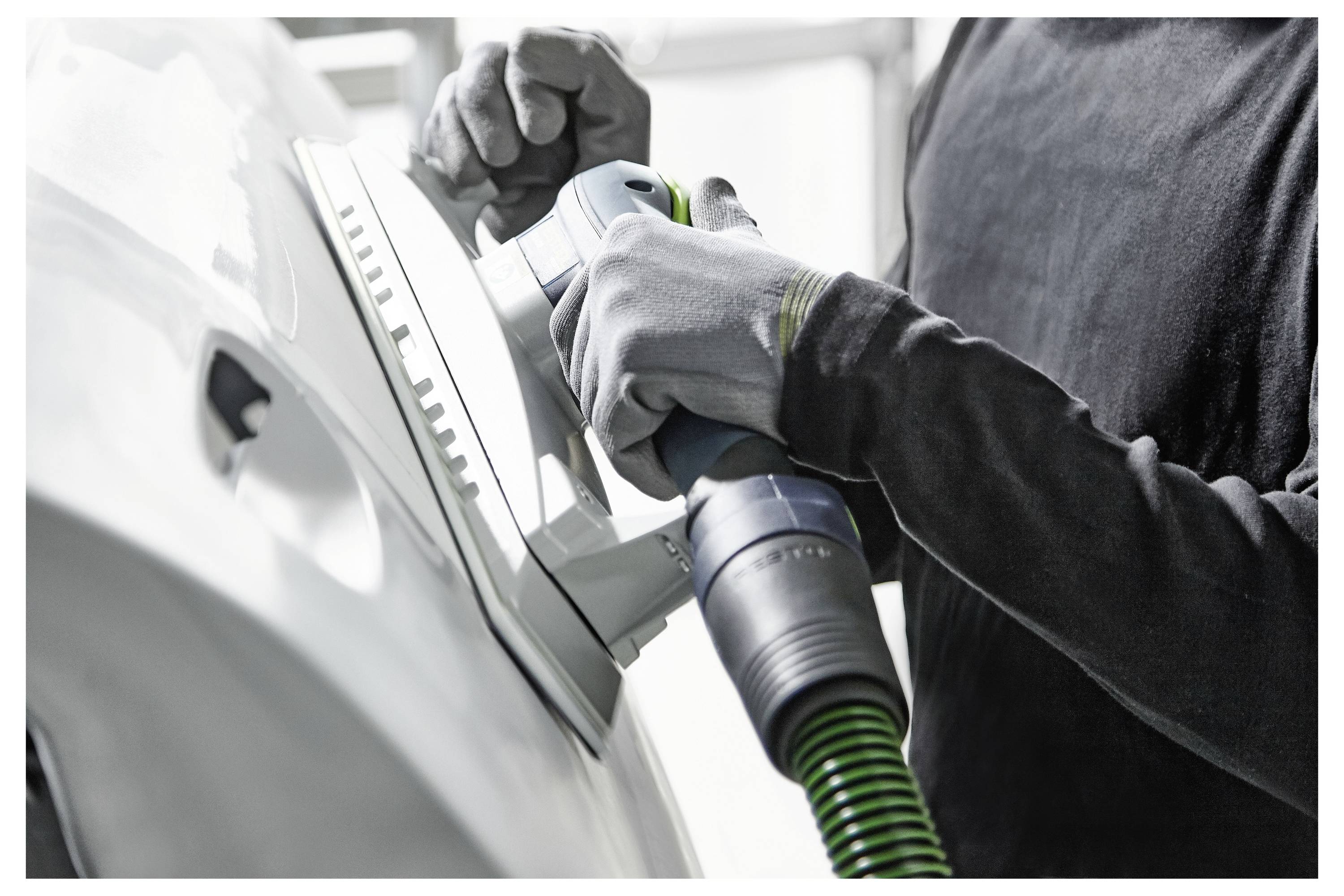 Eine Person mit Handschuhen, die einen Elektroschleiffer auf der Oberfläche eines Fahrzeugs verwendet, wahrscheinlich zum Polieren oder Glätten der Außenseite des Autos in einer Werkstatt.