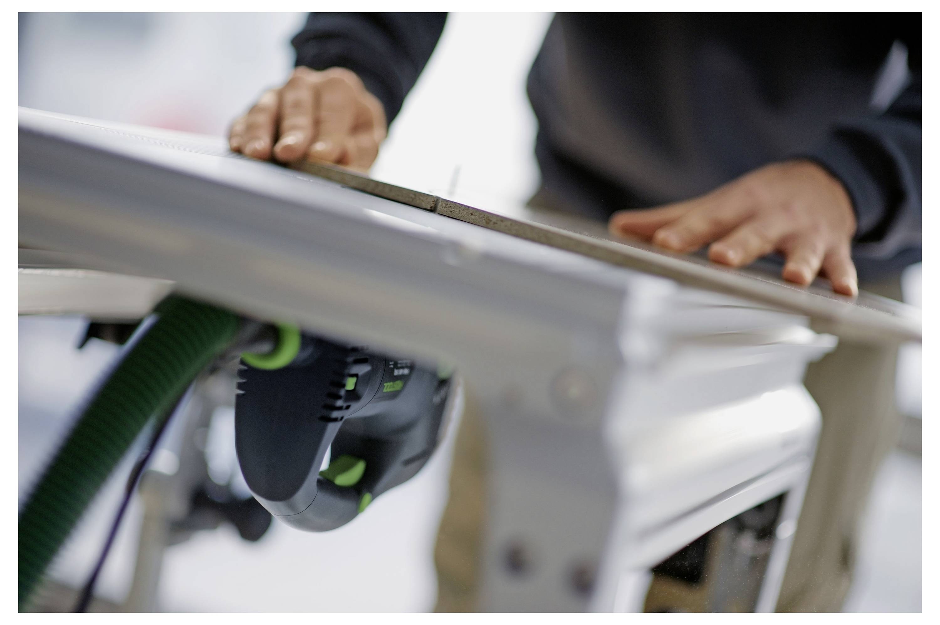 Eine Hand hält ein Festool-Elektrowerkzeug, das mit einem Schlauch ausgestattet ist, und demonstriert dessen Verwendung beim Durchtrennen eines Holzbretts und zeigt dabei Präzision.