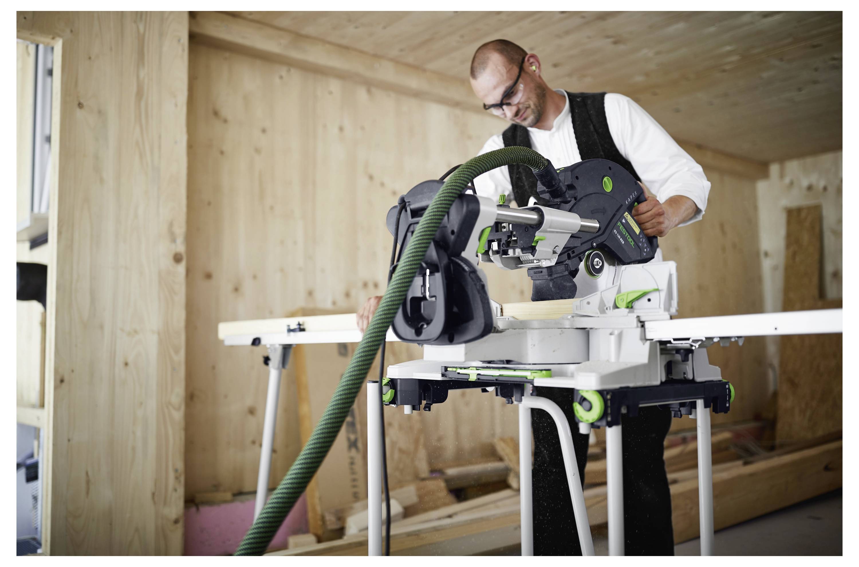 Eine Person benutzt eine Gehrungssäge in einer Holzwerkstatt. Die Säge hat einen grünen Staubsammelschlauch angebracht, und Holzbretter befinden sich in der Nähe.