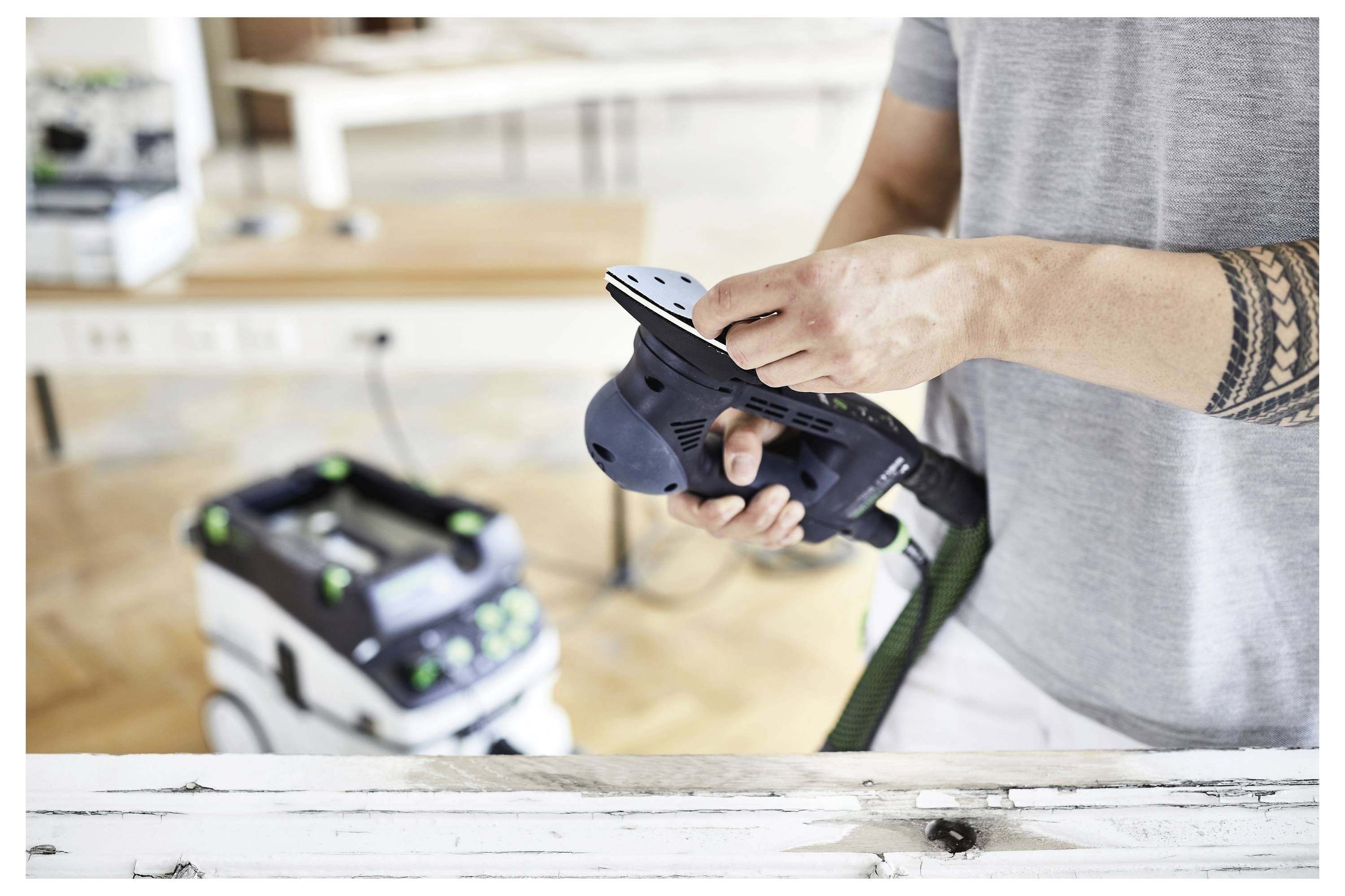 Eine Person wechselt das Schleifpapier an einem Elektroschleiffer in einer Holzwerkstatt, mit Geräten im Hintergrund sichtbar.